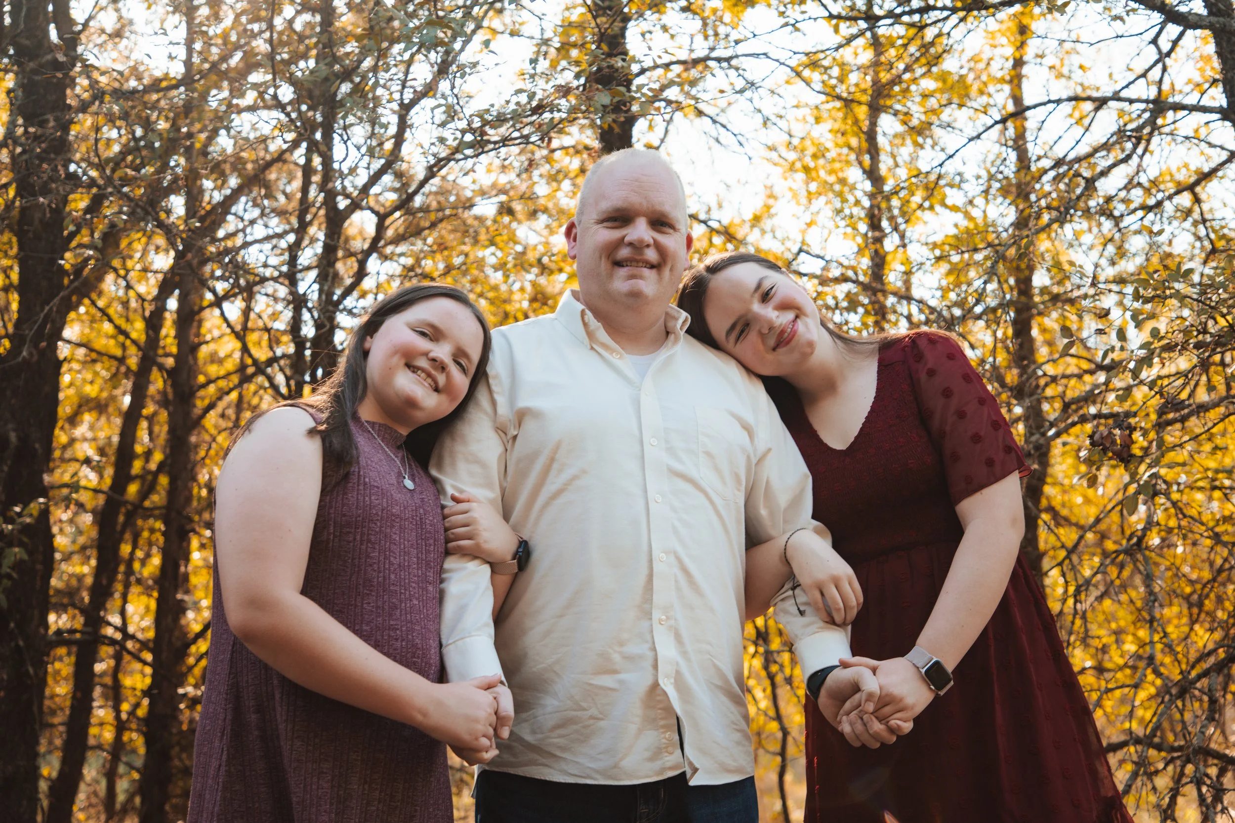 A family of three standing outdoors in a forest with autumn leaves, smiling and holding hands. The man is in the middle, wearing a cream-colored shirt, with a young girl on his left in a purple dress, and an older girl on his right in a burgundy dres