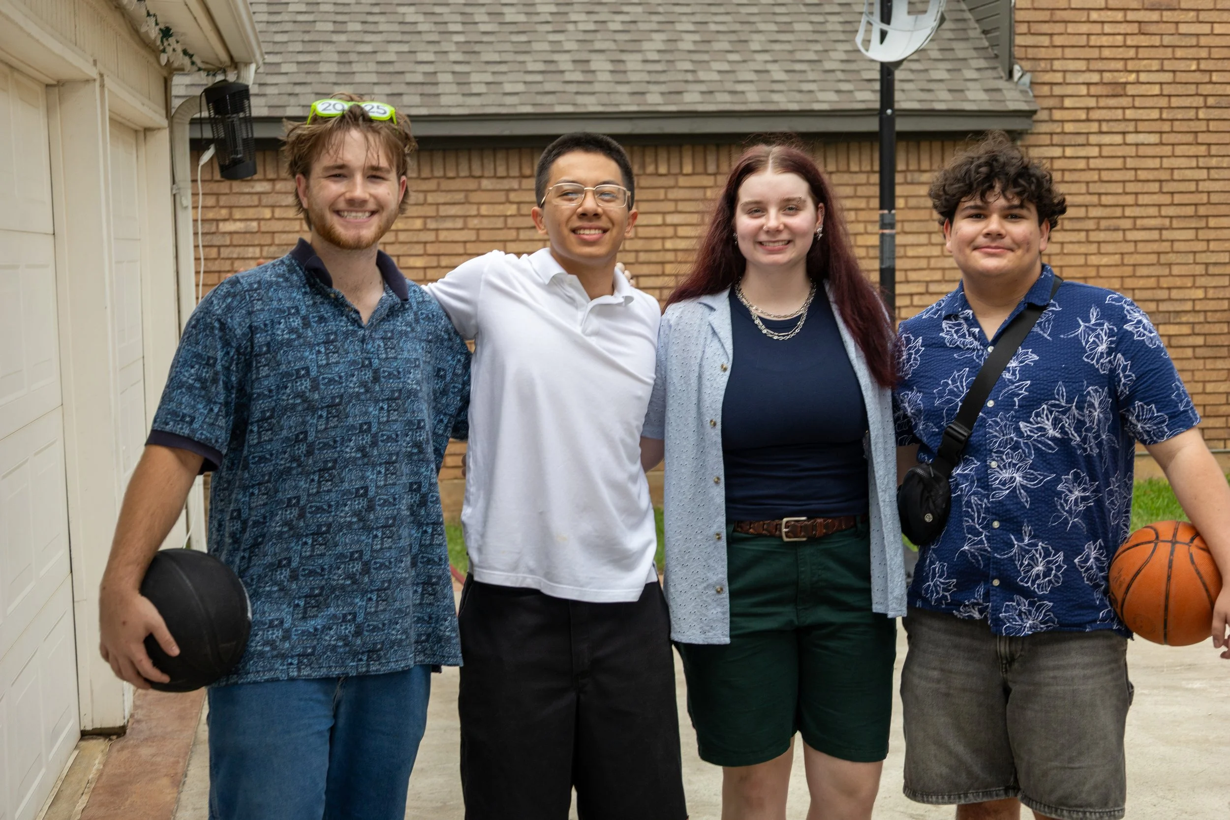 Four teenagers standing outdoors in a driveway, smiling at the camera. Two are holding basketballs, one with a camera strap across his chest, and one wearing glasses with a 2025 sticker on his glasses.