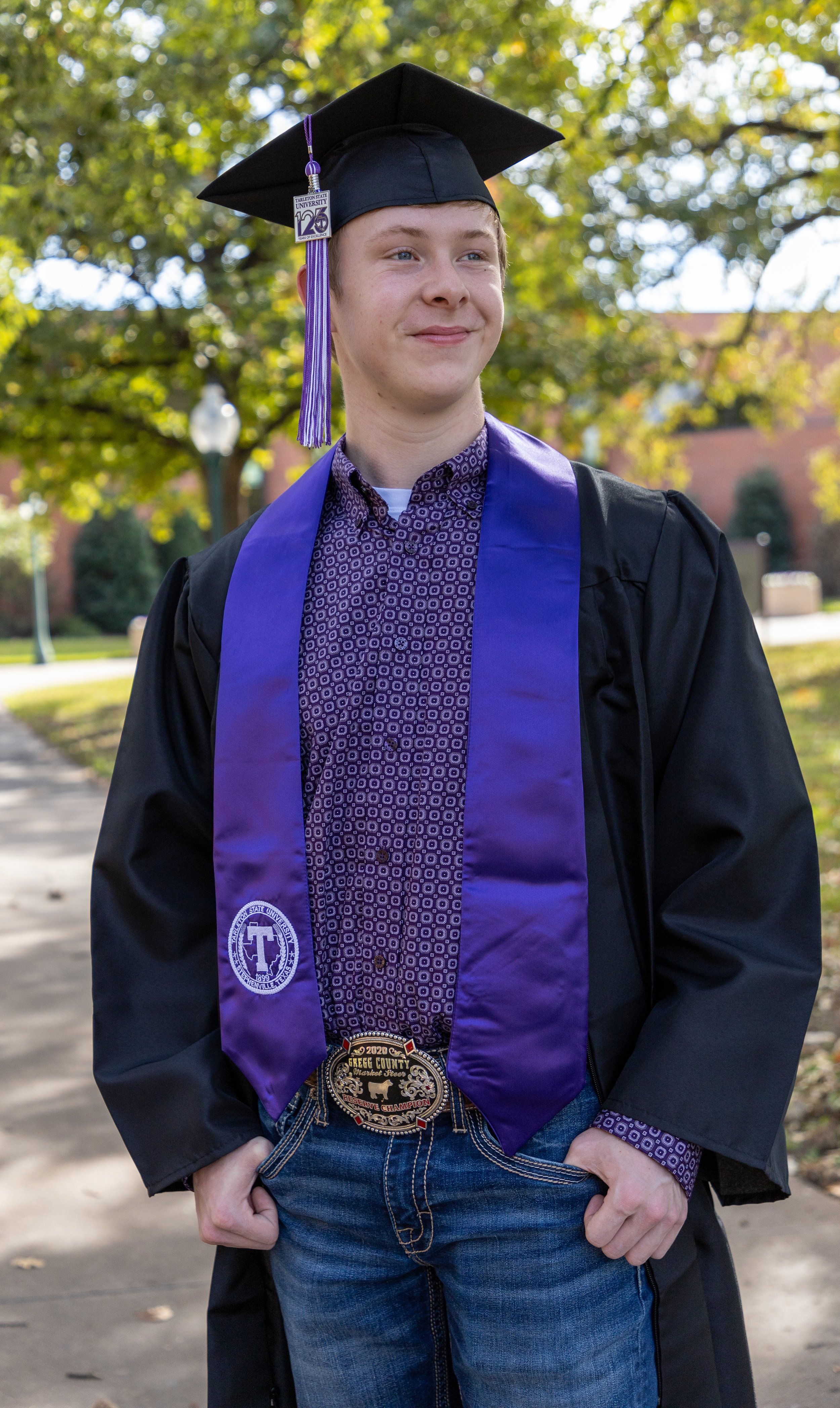 Young man in graduation cap and gown outdoors, wearing a purple stole and belt buckle, smiling.