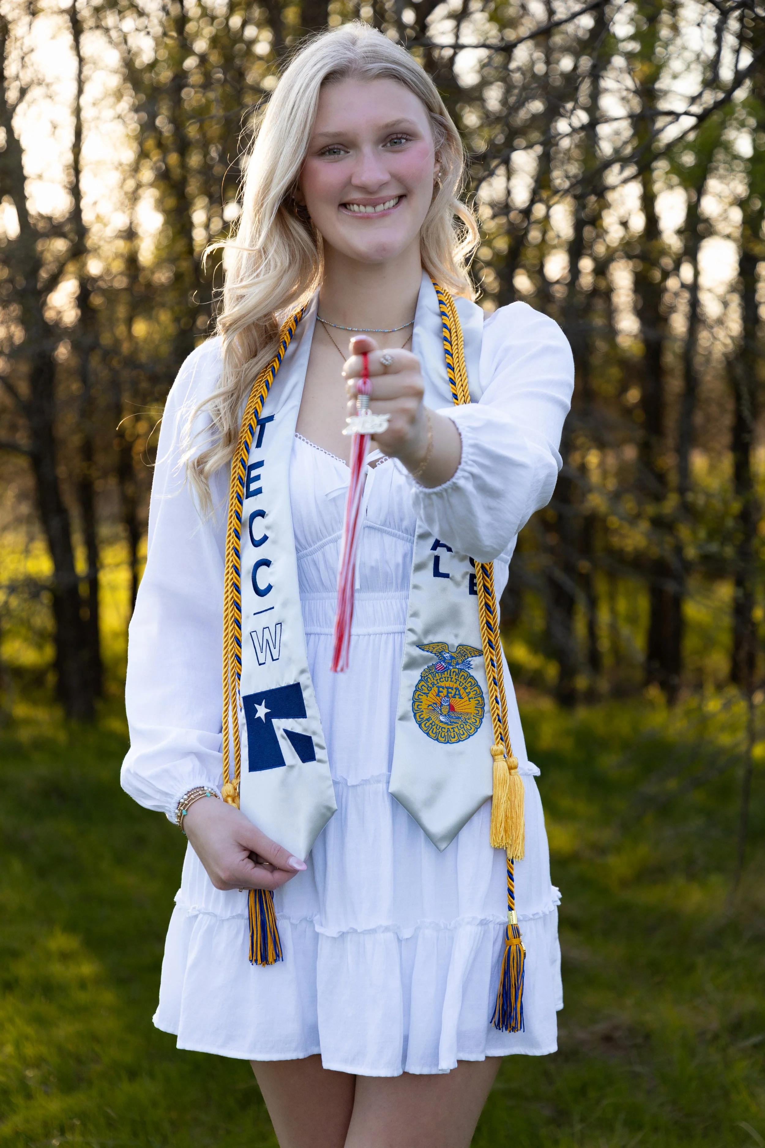 A young woman wearing a white dress and a graduation stole holds a diploma outside, with trees in the background.