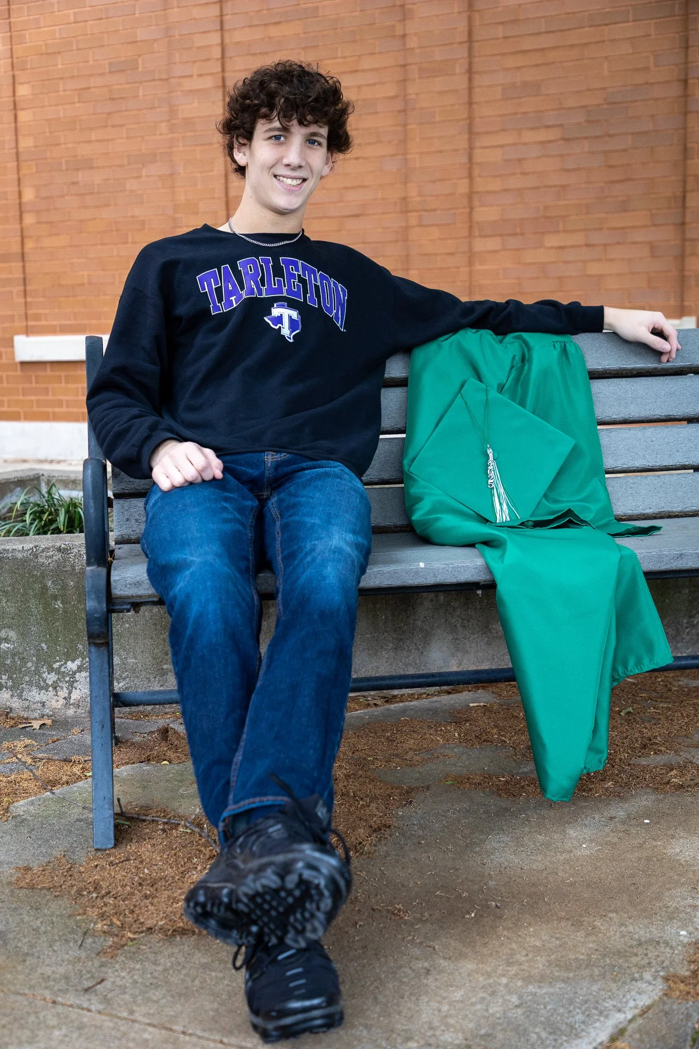 A young man with curly hair sitting on a park bench, wearing a black Tarleton sweatshirt, jeans, and black shoes. A green graduation gown and cap are draped over the bench beside him.