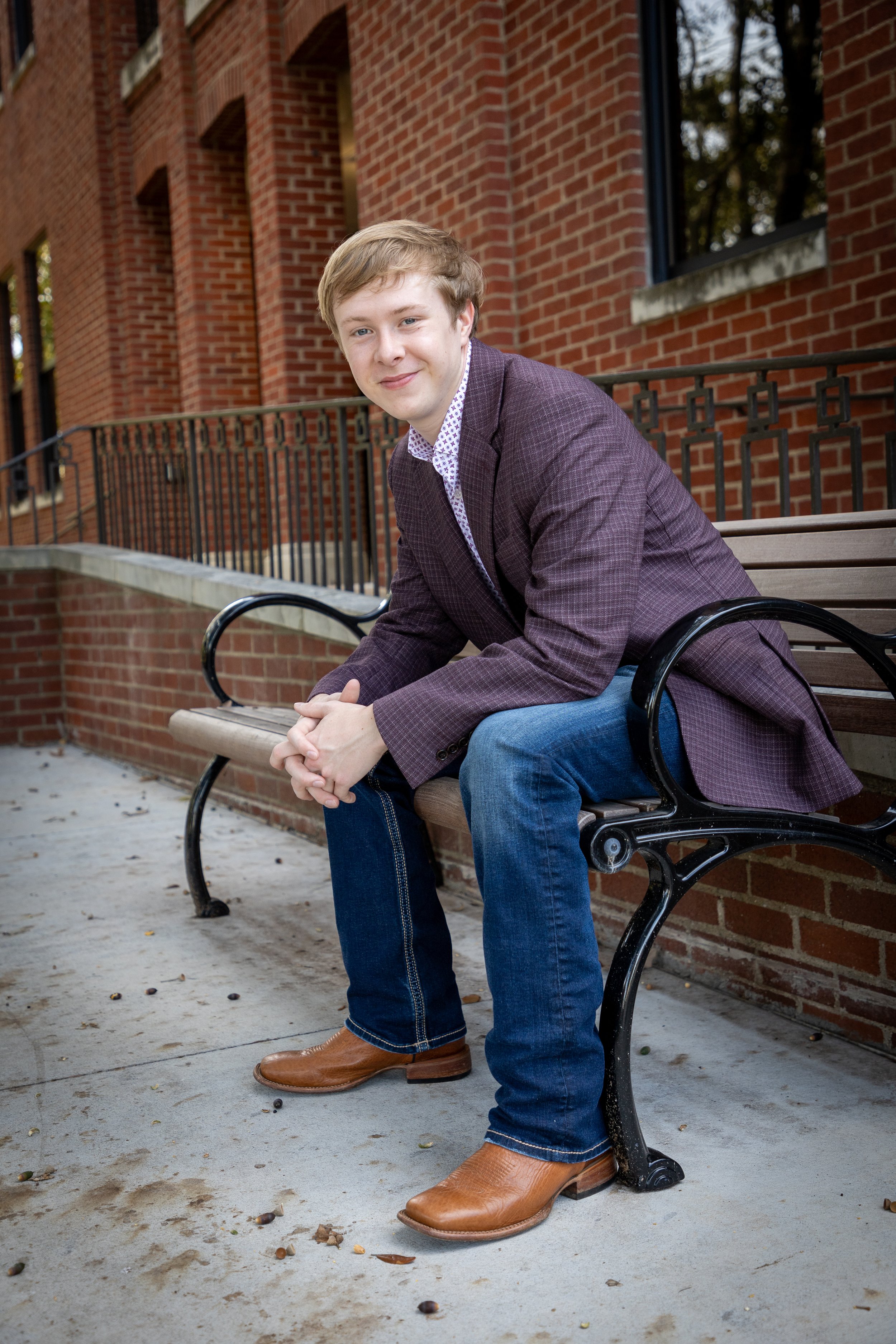 Young man sitting on a park bench in front of a brick building, wearing a blazer, jeans, and brown shoes, smiling at the camera.