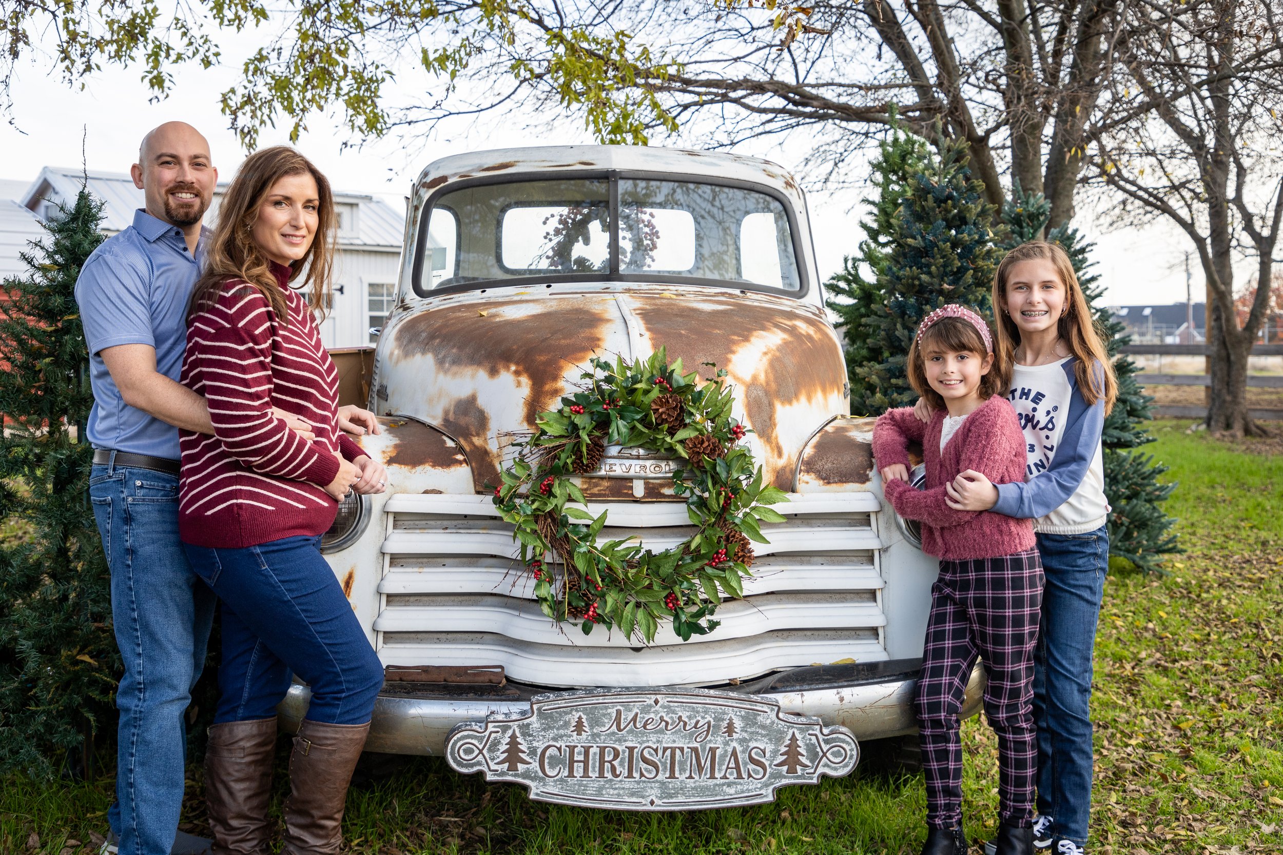 Family of four standing beside a vintage rusted Chevrolet truck decorated with Christmas wreath and signs, Christmas trees in the background, outdoors on a fall day.