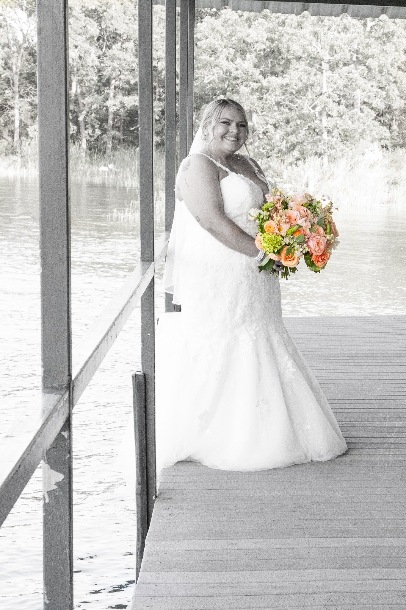 A smiling bride in a lace wedding dress holding a colorful bouquet of peach, pink, and green flowers on a wooden dock beside a body of water with trees in the background.