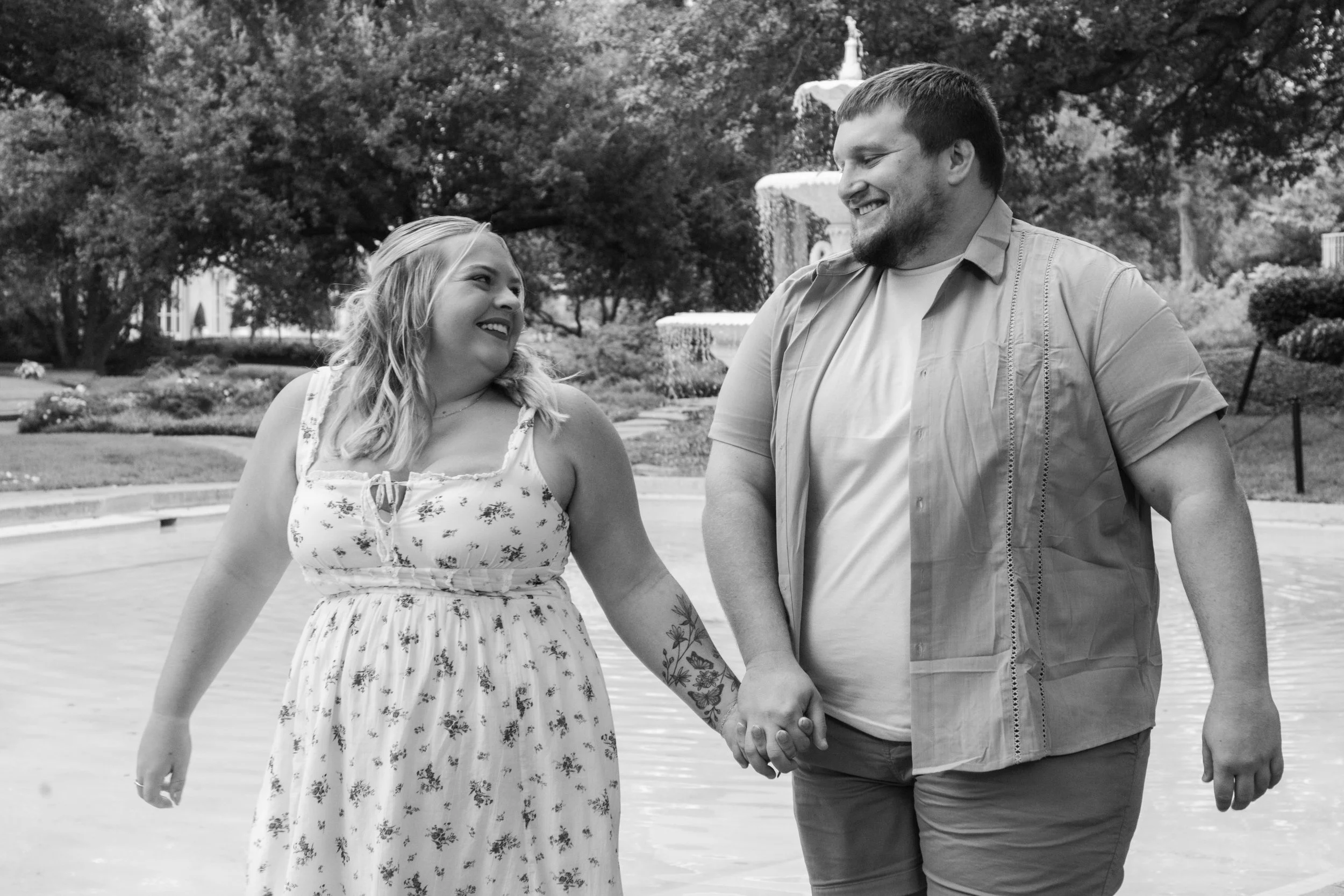 A smiling couple holding hands and walking near a water fountain in a park.