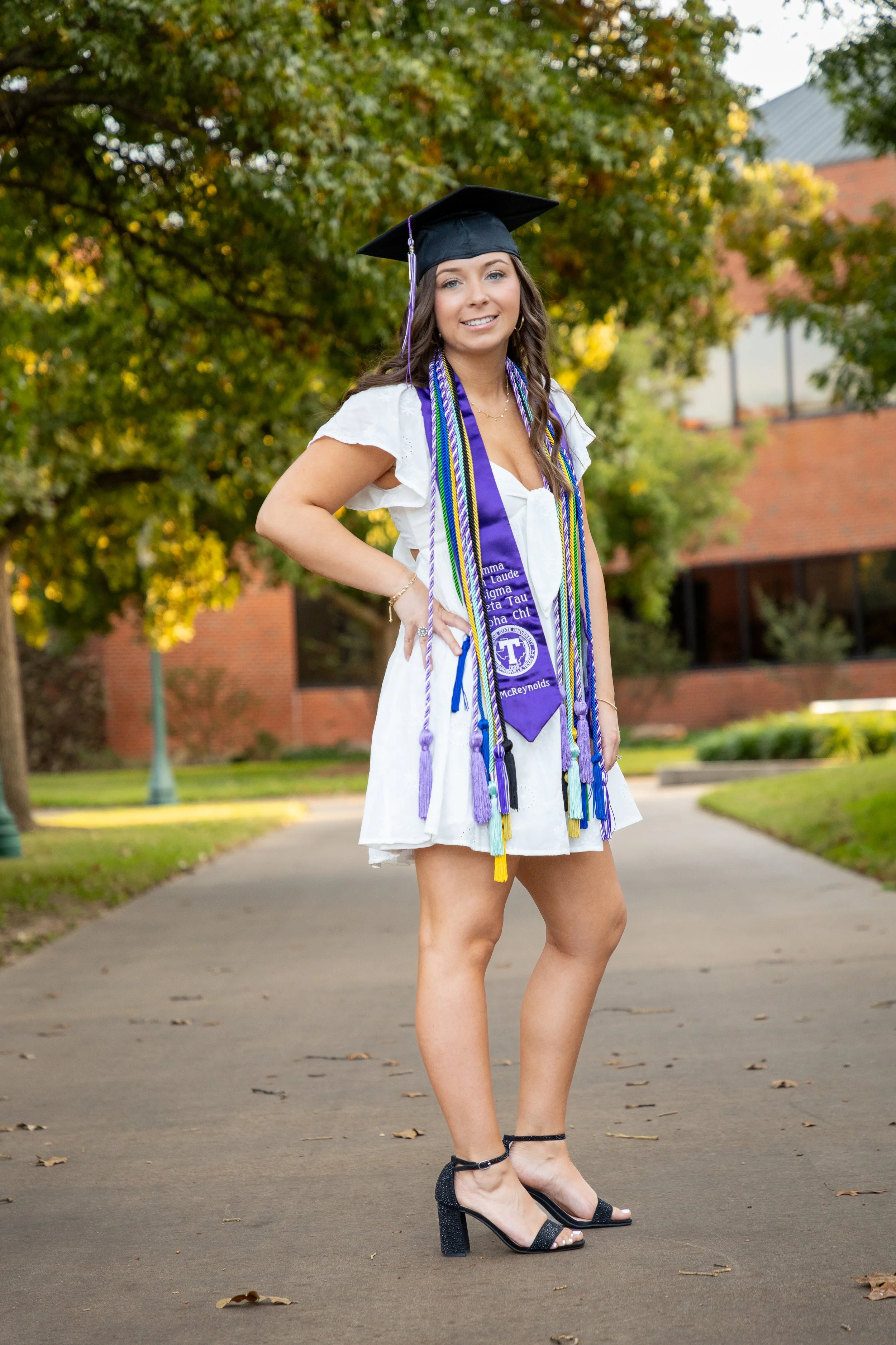 A young woman in a graduation cap and white dress standing outdoors on a sidewalk, smiling with her hands on her hips, with trees and a brick building in the background.