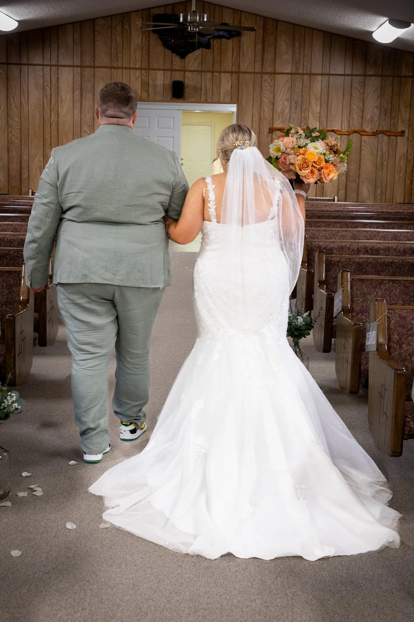 A bride and groom walking down the aisle in a church. The bride wears a white wedding gown with a veil, and the groom is in a gray suit. The bride holds a bouquet of orange and pink flowers.