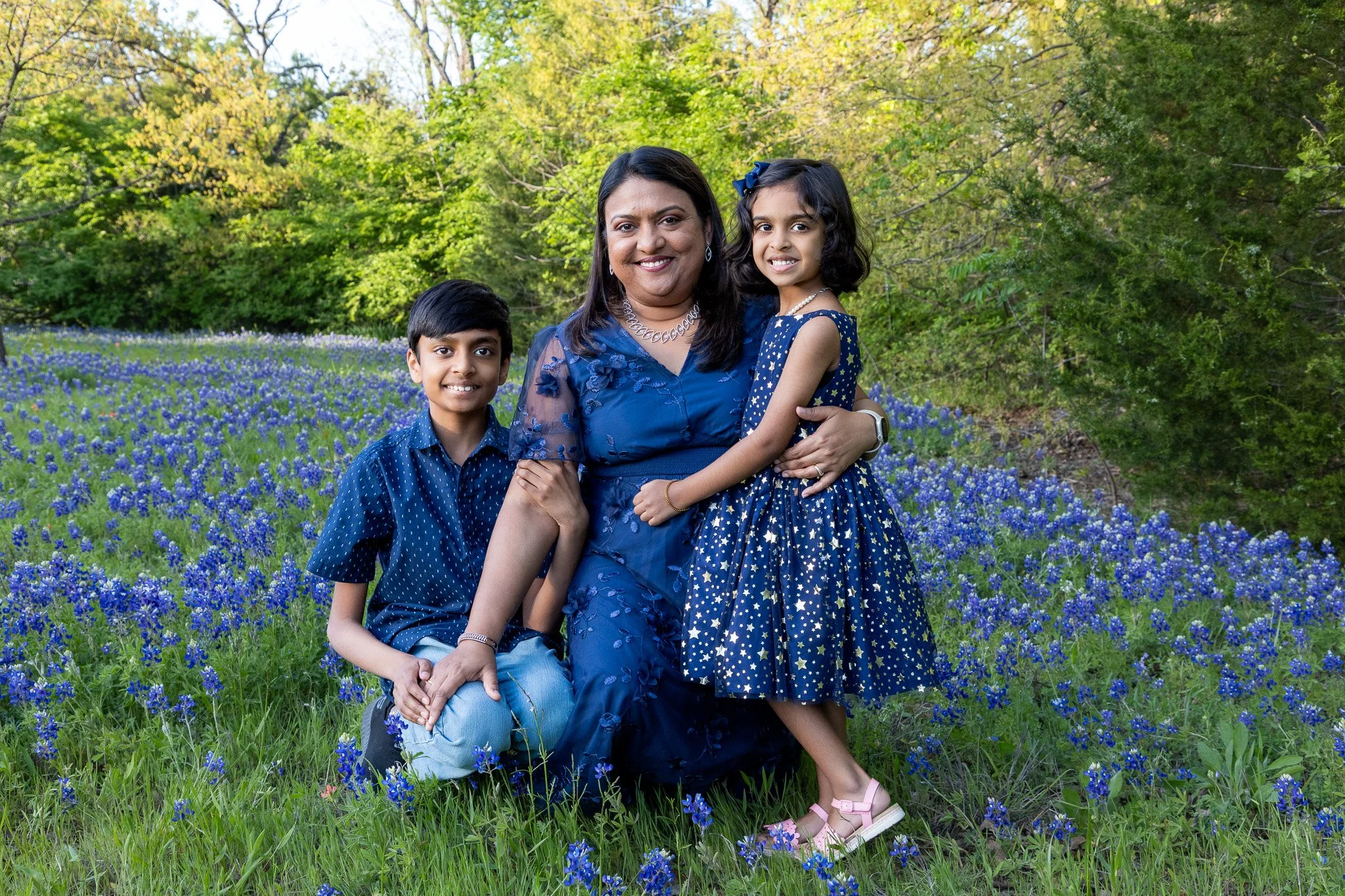A family of three standing in a field of blue flowers with trees in the background. The woman is in the center, wearing a blue dress, holding a young girl in a navy dress with star patterns. The boy is kneeling with one knee on the ground, wearing a 