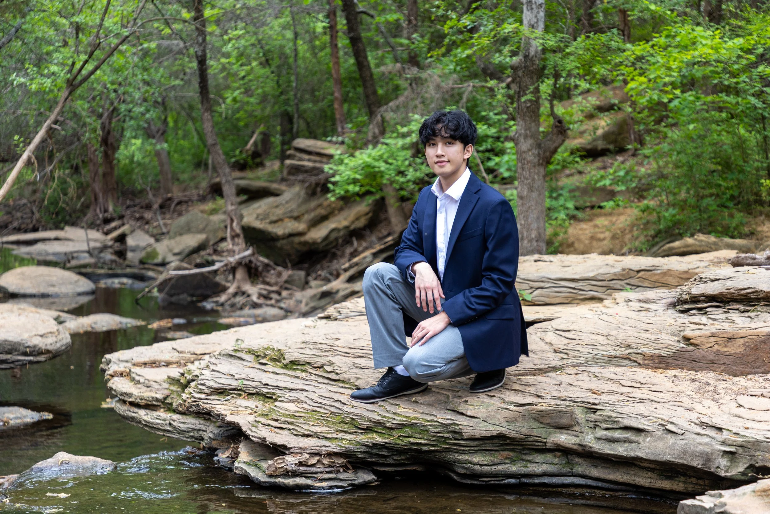 A young man in a suit kneeling on a large flat rock by a creek in a wooded area.