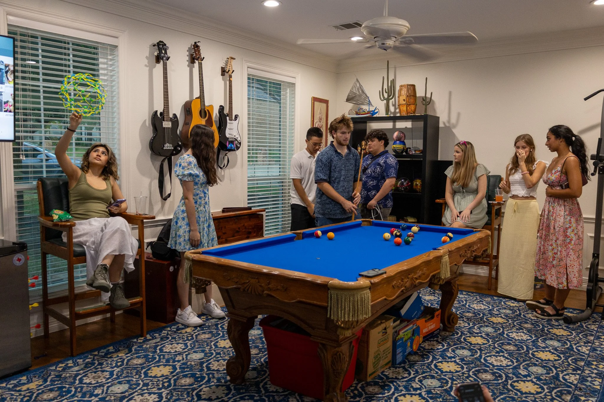 Group of young people in a living room around a pool table, some playing billiards, others watching and socializing, with guitars hanging on the wall and decorative items.