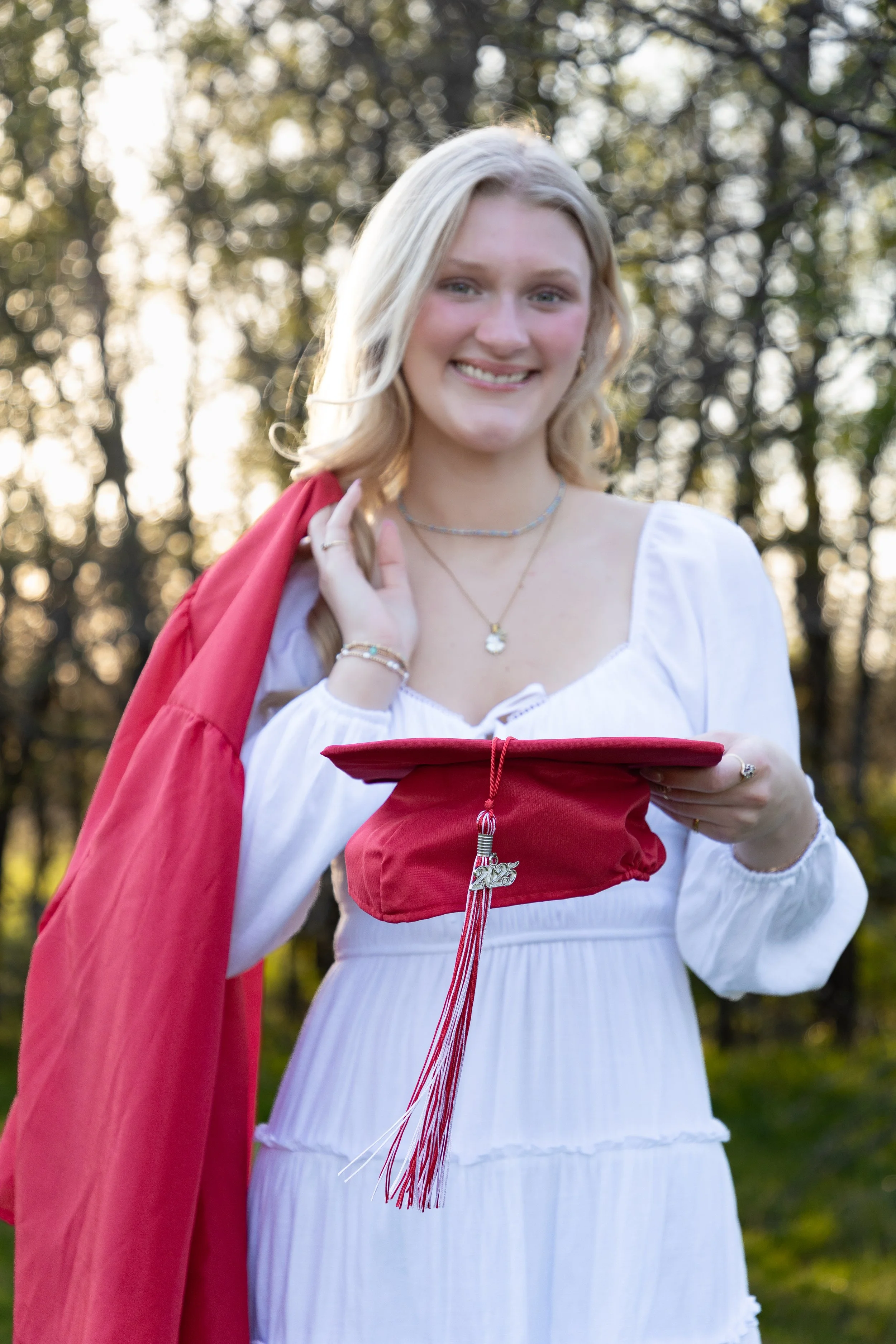 A young woman in a white dress holding a red graduation cap with a tassel and a charm, standing outdoors with trees in the background, smiling at the camera.