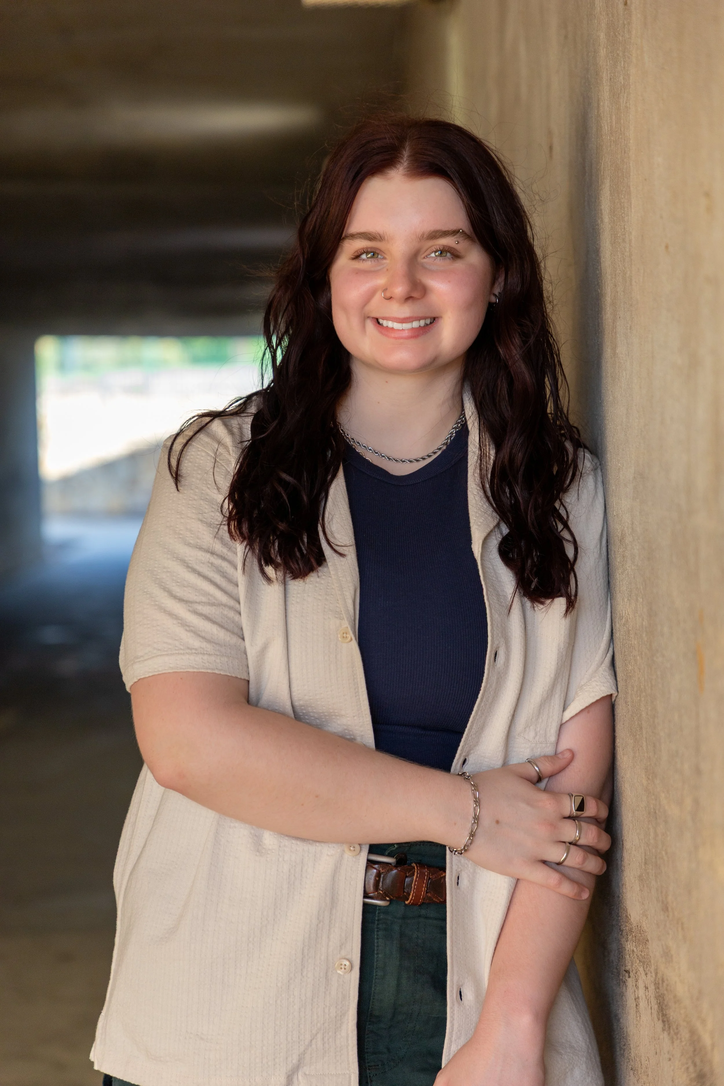 A young woman with brown, curly hair wearing a black shirt, beige open shirt, green pants, and jewelry, smiling and leaning against a yellow wall.