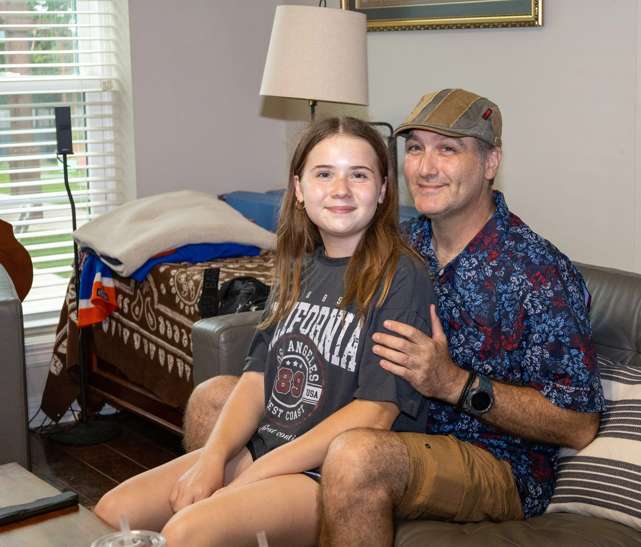 A girl and a man sitting on a couch in a living room, smiling at the camera. The girl has long red hair, and the man is wearing a cap and a floral shirt.