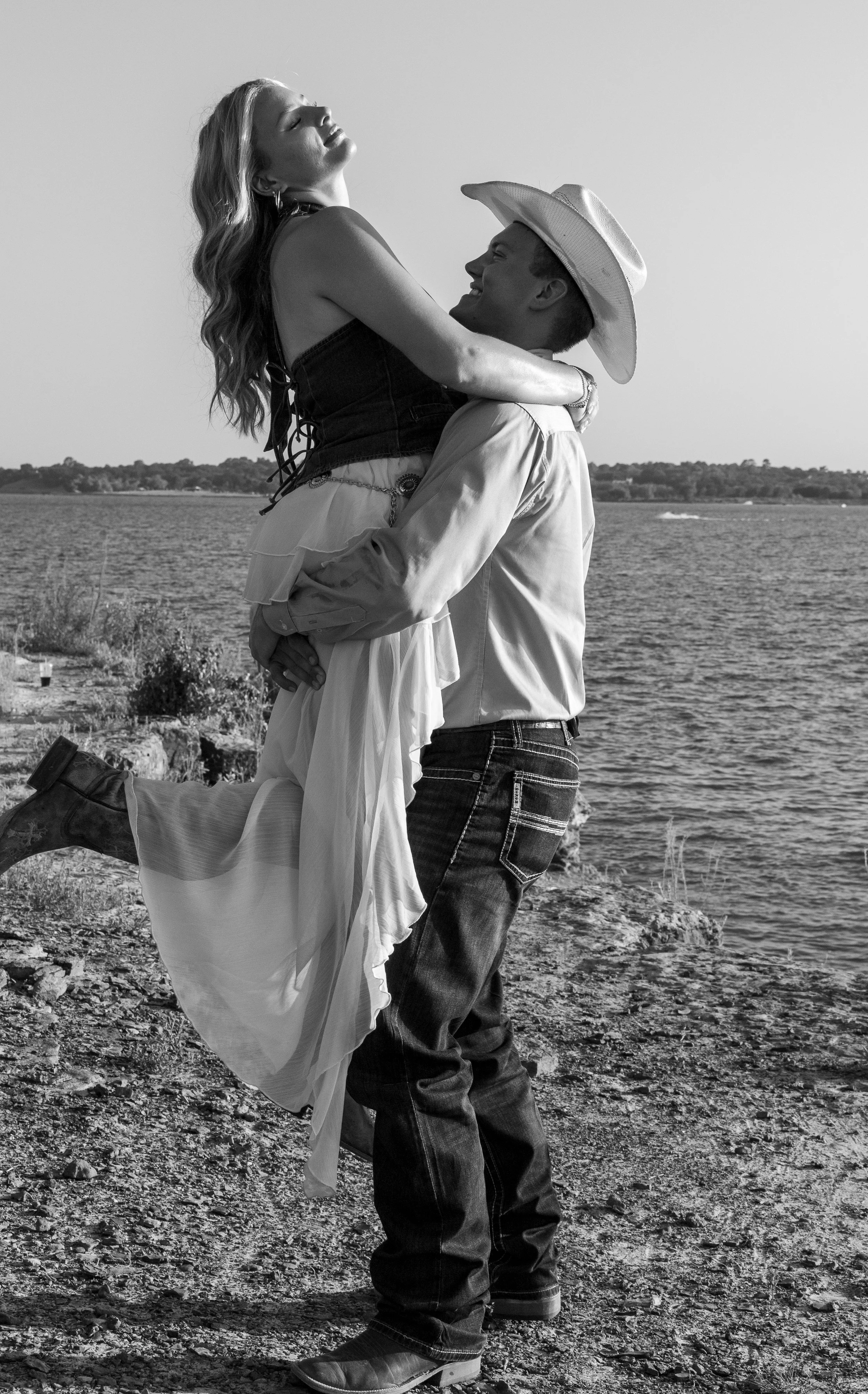 A black and white photo of a couple by a lake, with the woman being lifted by the man. The woman has long wavy hair, is wearing a sleeveless top and a flowing skirt, and appears relaxed with her eyes closed. The man is wearing a cowboy hat, a long-sl