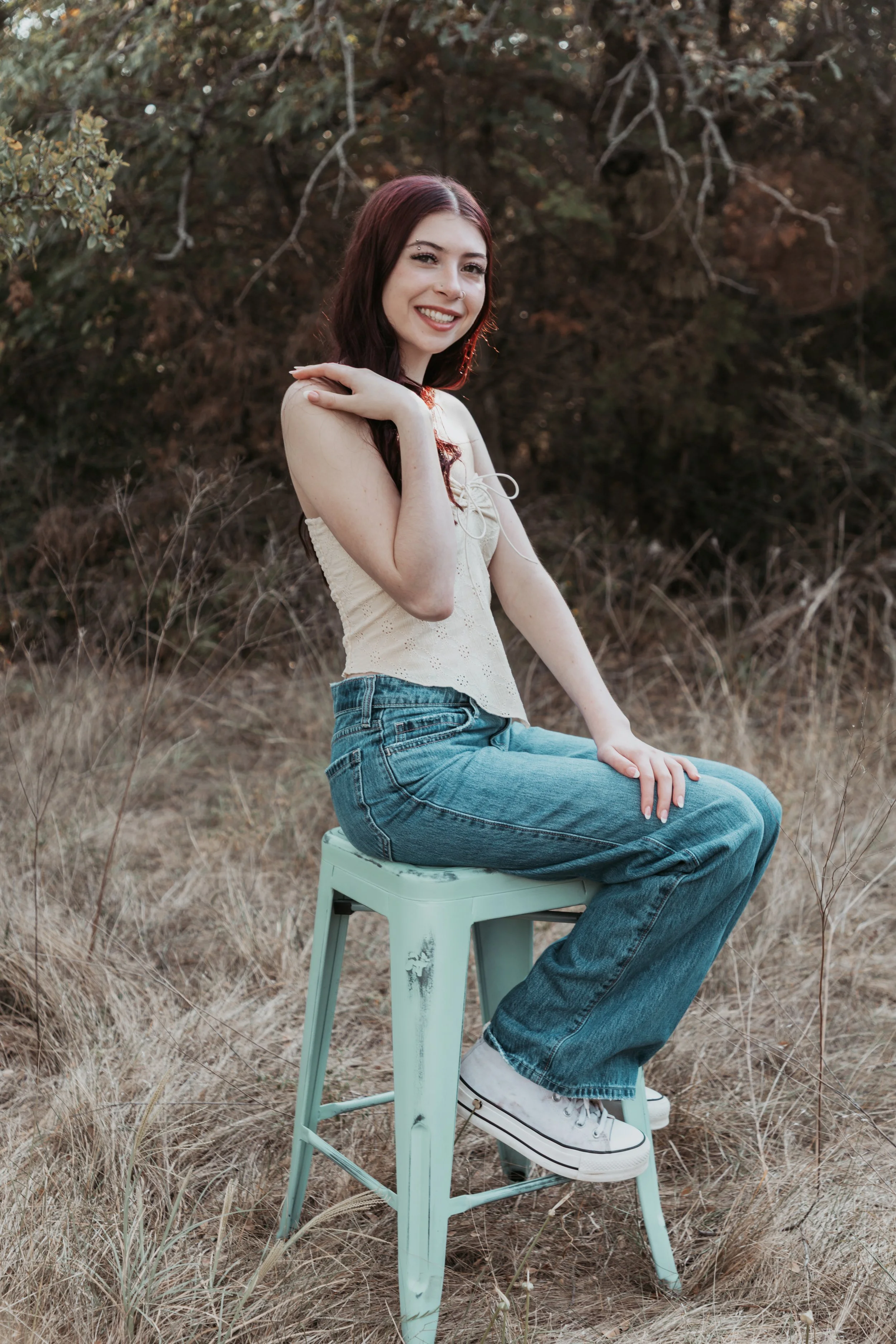A young woman with long dark red hair sitting on a light green metal chair outdoors in a field with dry grass and bushes, smiling at the camera.