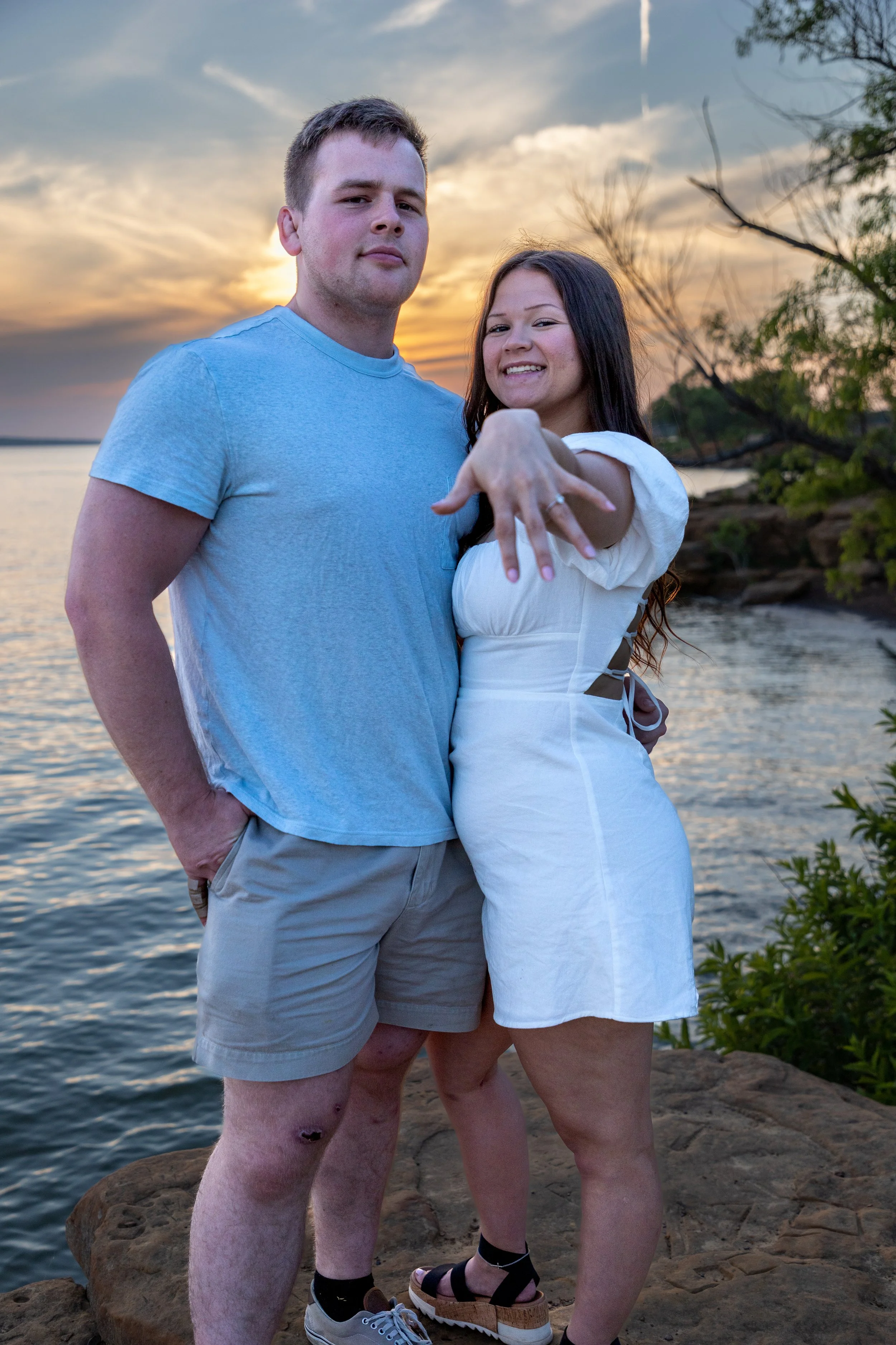 A young couple standing on rocks by a body of water at sunset, with one woman showing off her engagement ring and smiling.