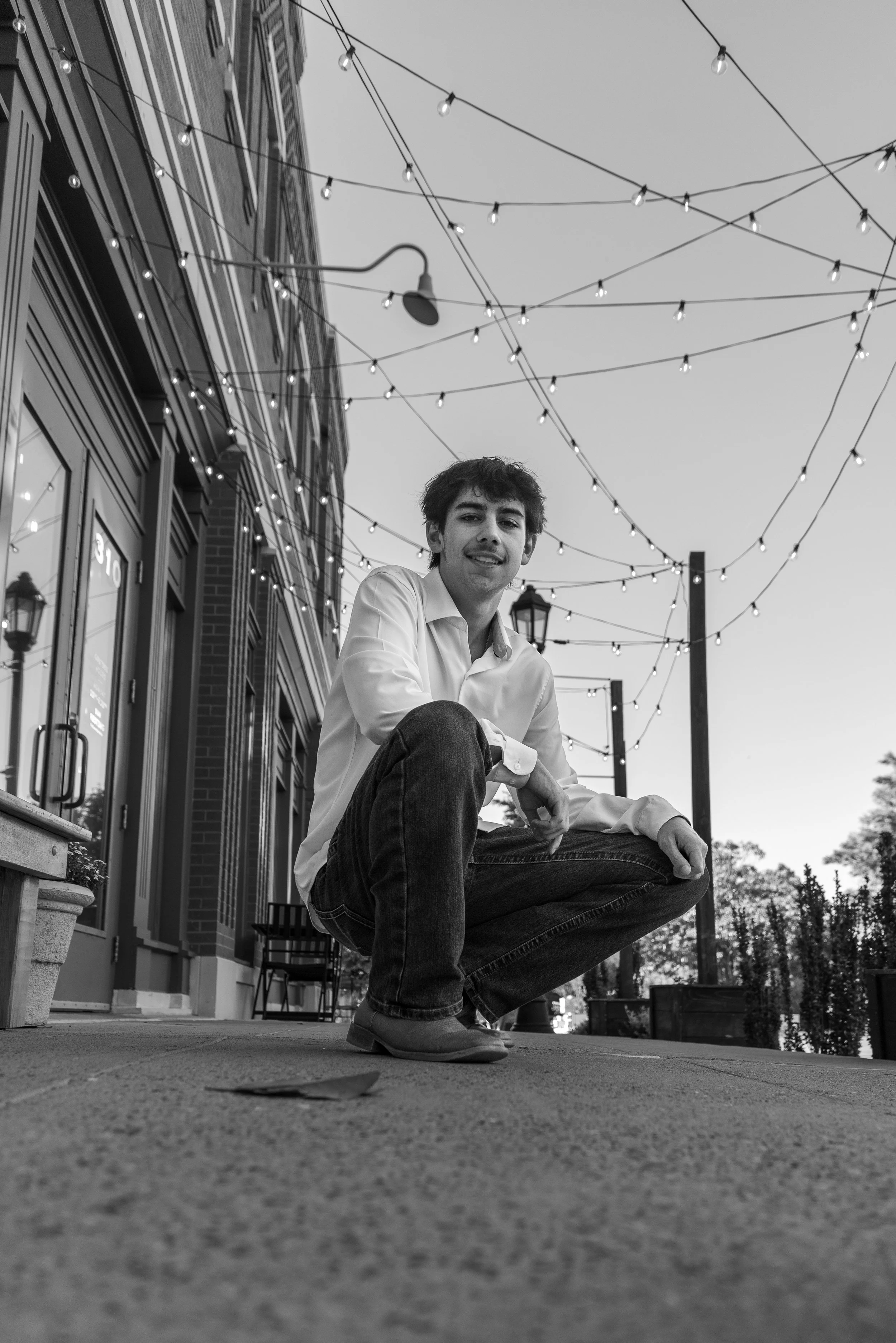 A young man crouching on a sidewalk outdoors during the evening, with string lights overhead and a building facade with large windows behind him.