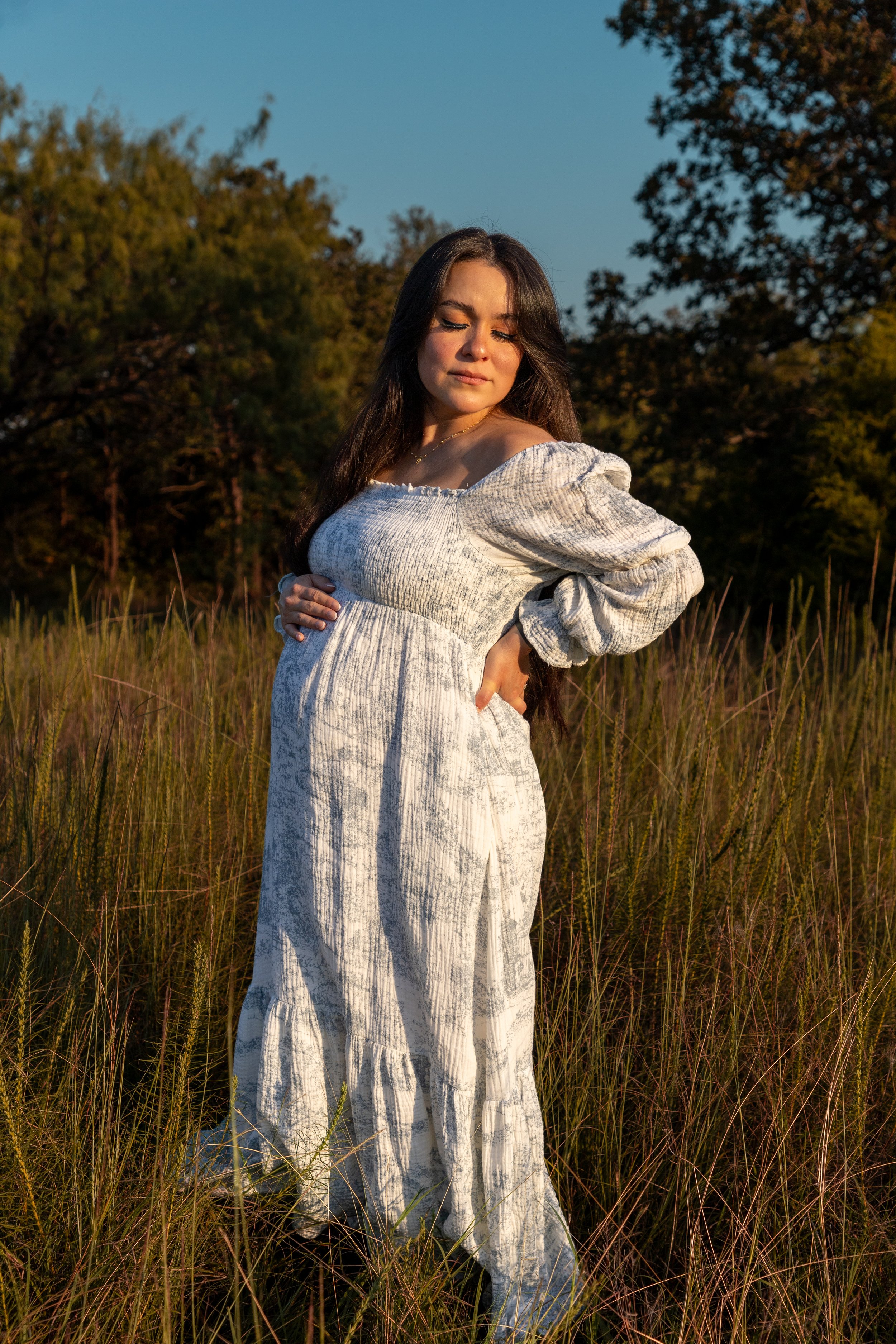 A pregnant woman in a white dress standing in a grassy field during golden hour sunset.