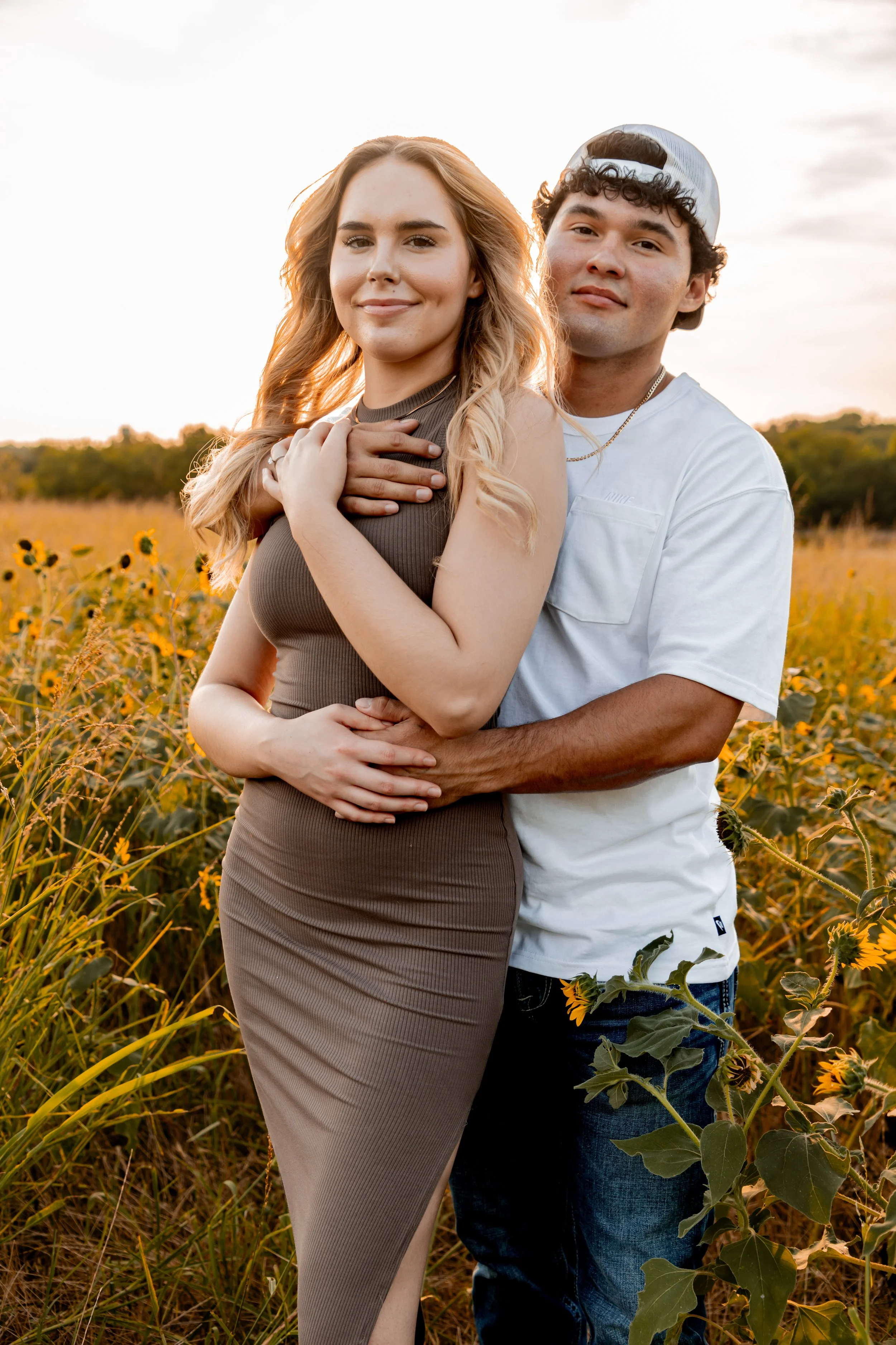A smiling young woman in a beige dress stands in a sunflower field at sunset while a young man in a white t-shirt and baseball cap stands behind her, both embracing each other.