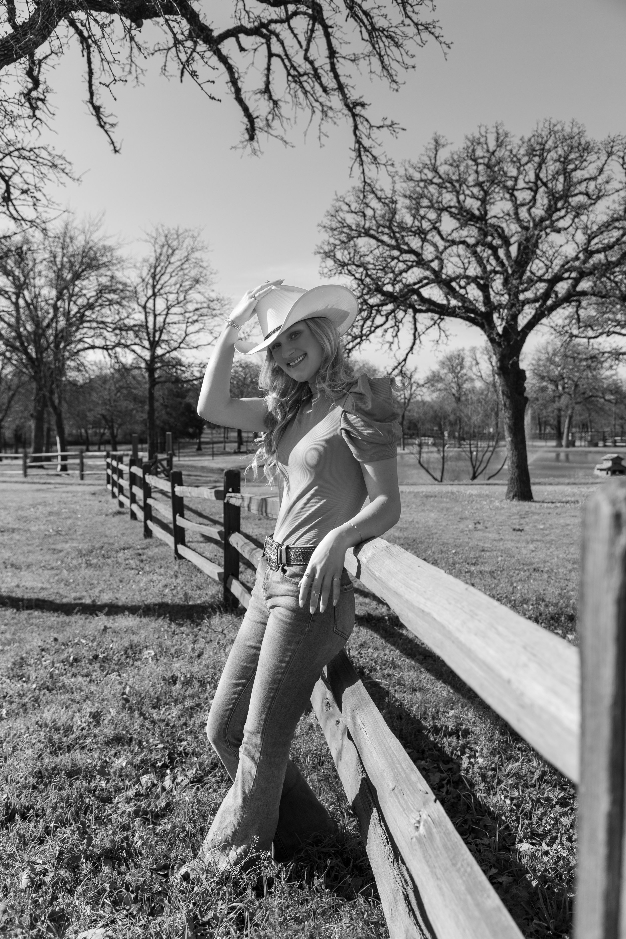 A woman wearing a cowboy hat and casual clothes leaning on a wooden fence in a park with leafless trees and open grassy areas.