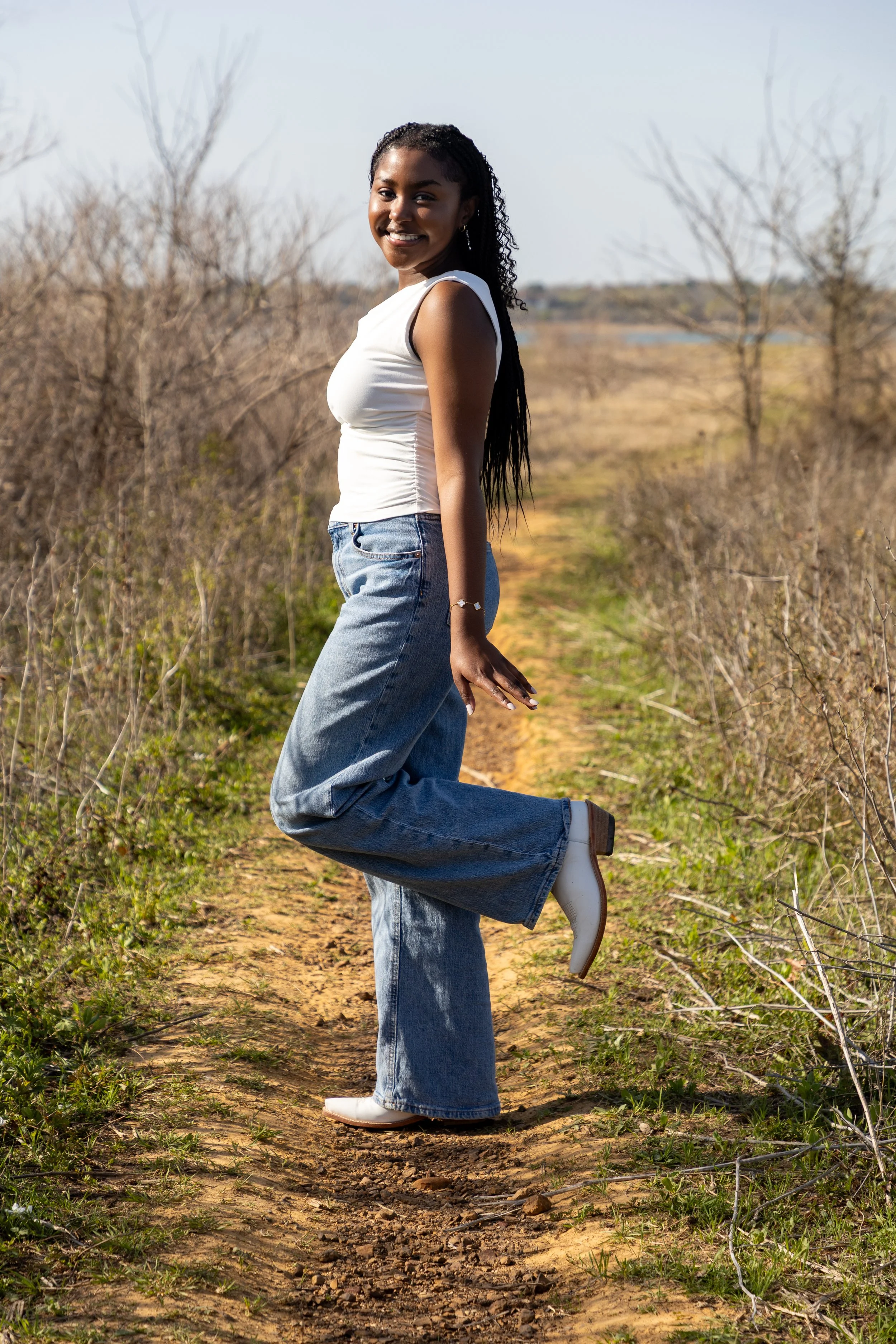 A smiling woman standing on a dirt path in a dry, open field during the daytime, wearing a white sleeveless top, blue jeans, and white boots.