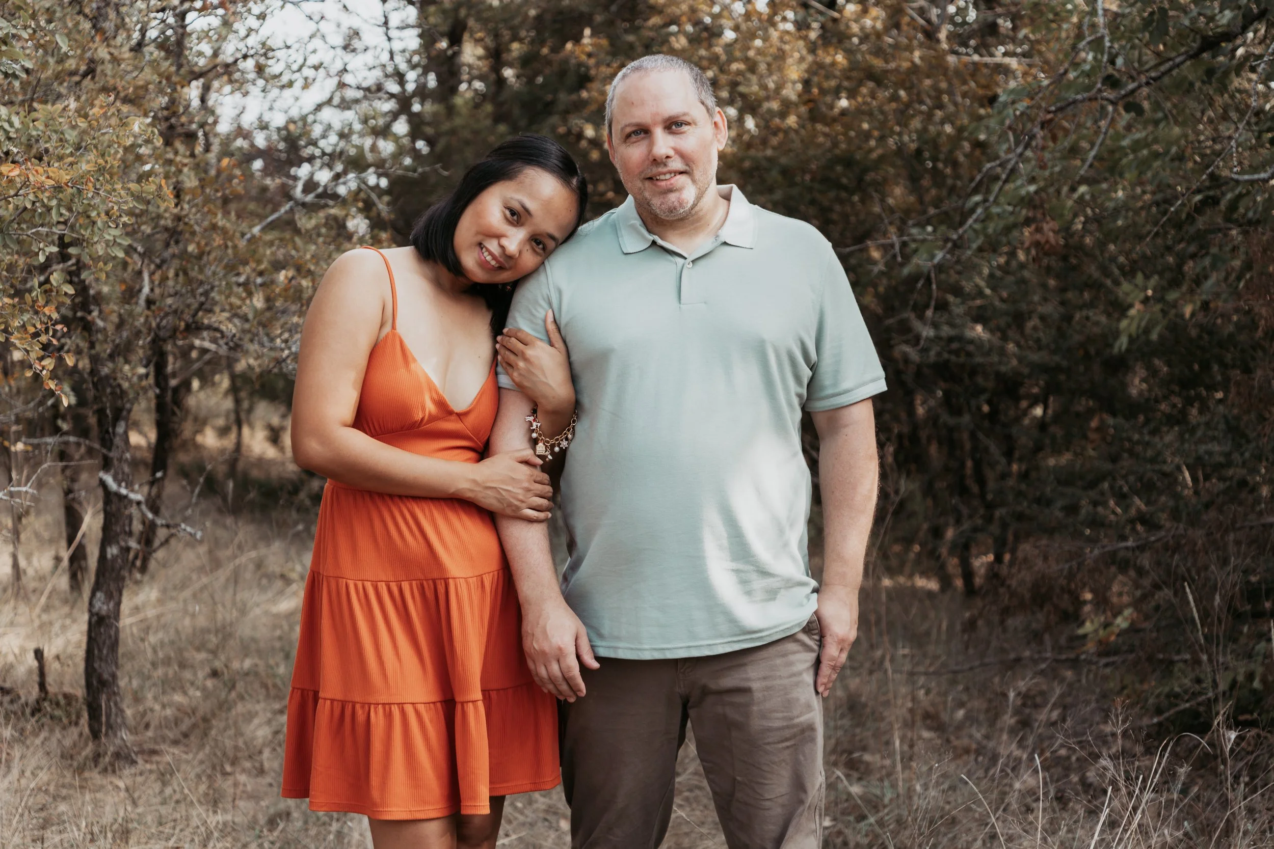 A woman in an orange dress leaning on a man in a light green polo shirt, standing outdoors in front of trees with autumn leaves.