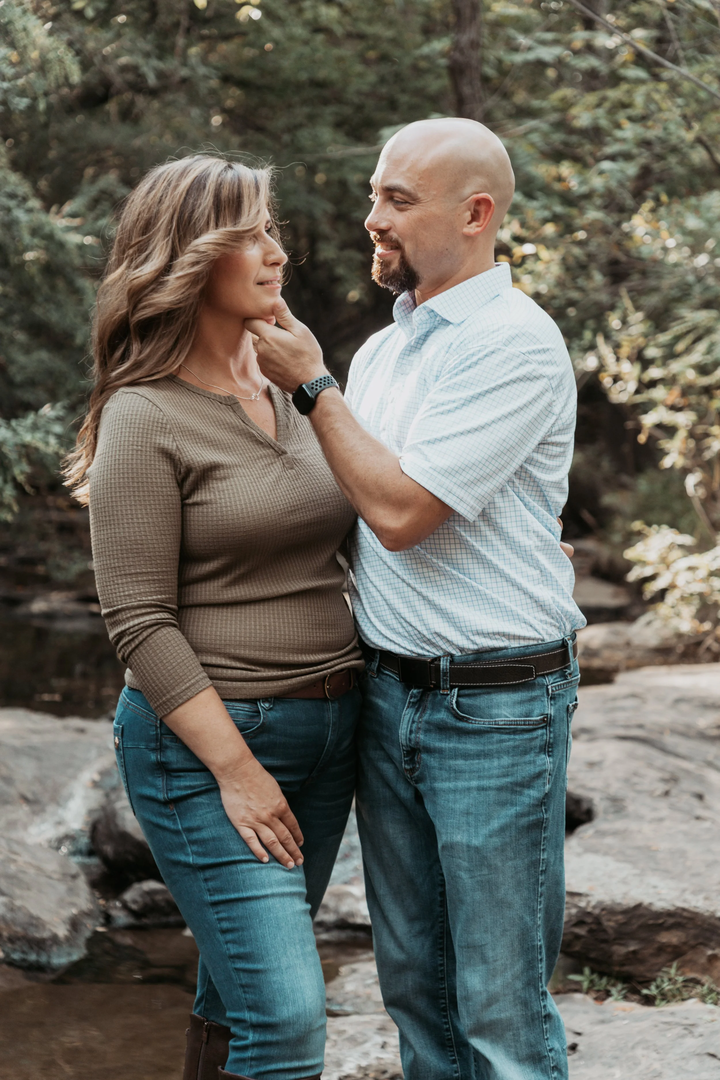 A couple standing close together outdoors in a wooded area, gazing into each other's eyes, with the man touching the woman's chin gently.