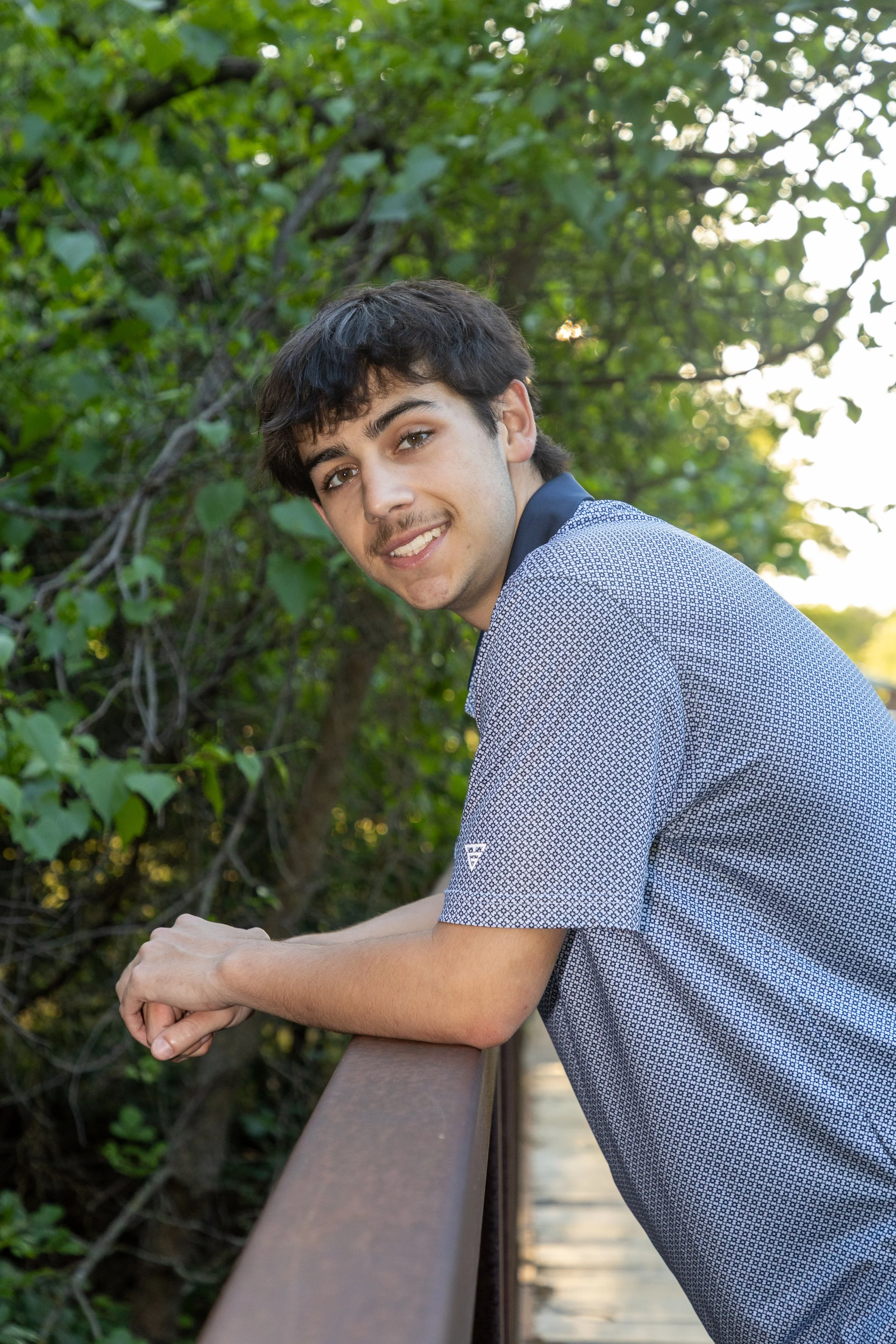 A young man with dark hair and light skin leaning on a wooden railing outdoors, smiling at the camera, with green foliage and trees in the background during daytime.