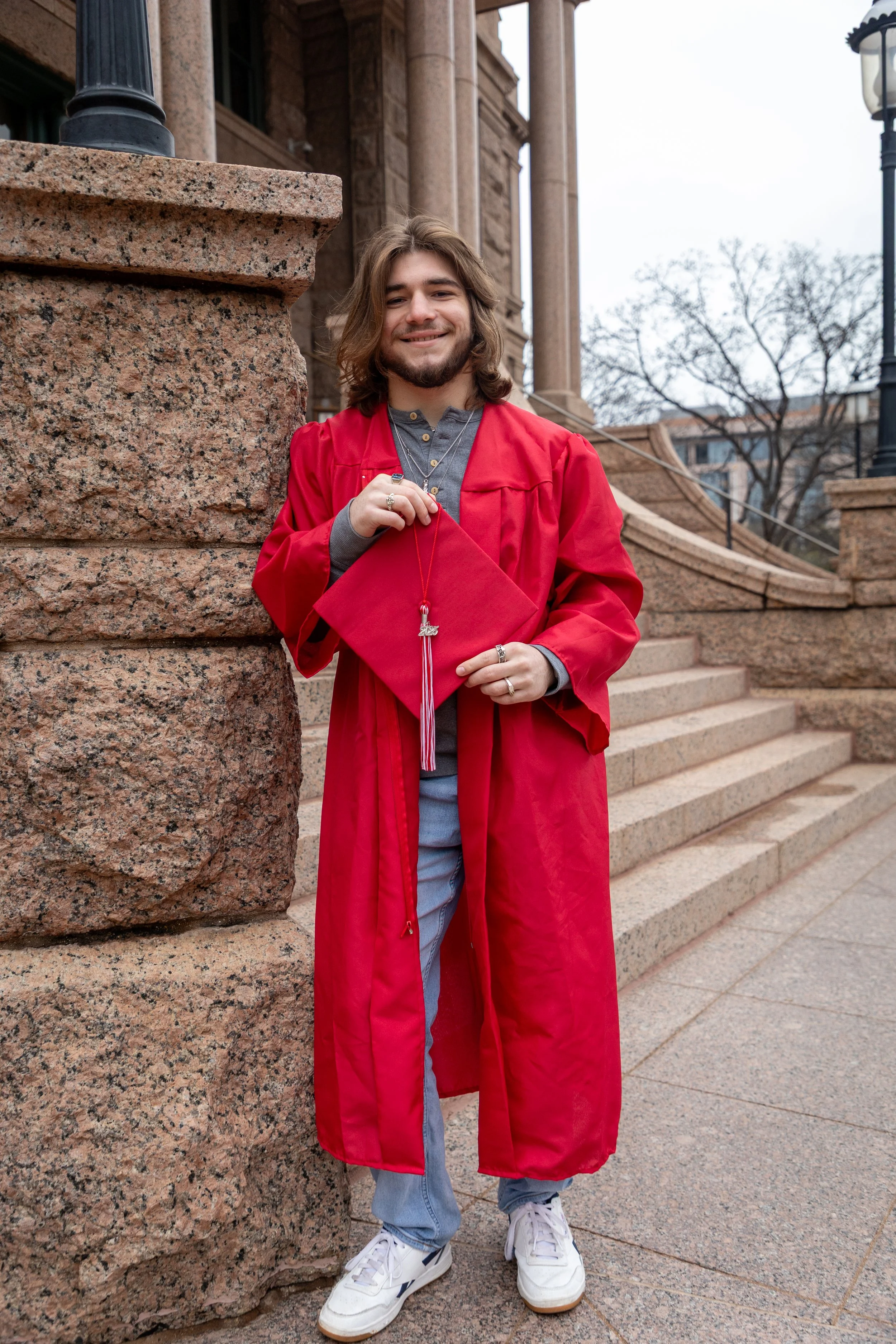 A young man in a red graduation gown and holding a matching cap, standing outside on steps near a large stone building.