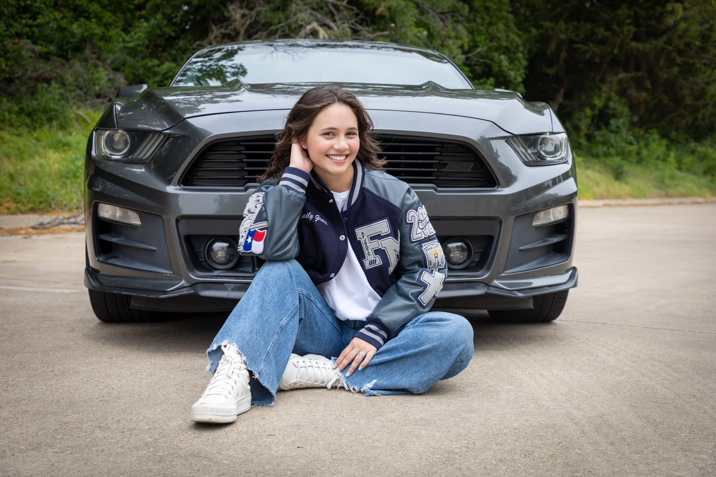 Young woman sitting on the ground in front of a gray sports car, smiling at the camera, wearing a blue varsity jacket and jeans.
