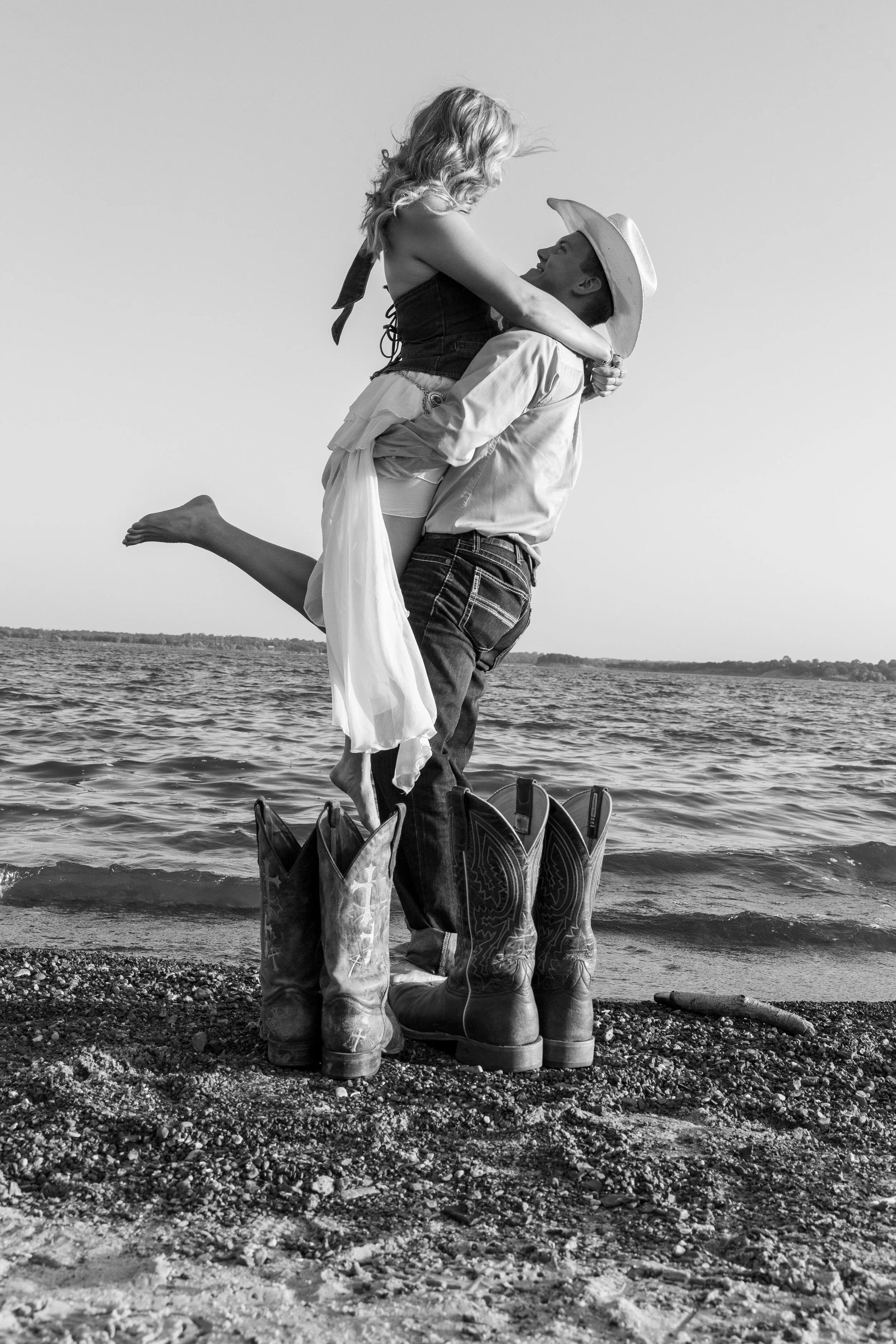 A couple stands by a lake, with the man holding the woman in his arms as she leans into him. They are dressed casually, and the woman has her legs wrapped around the man. The scene is outdoors on a rocky beach, with cowboy boots at their feet and a c