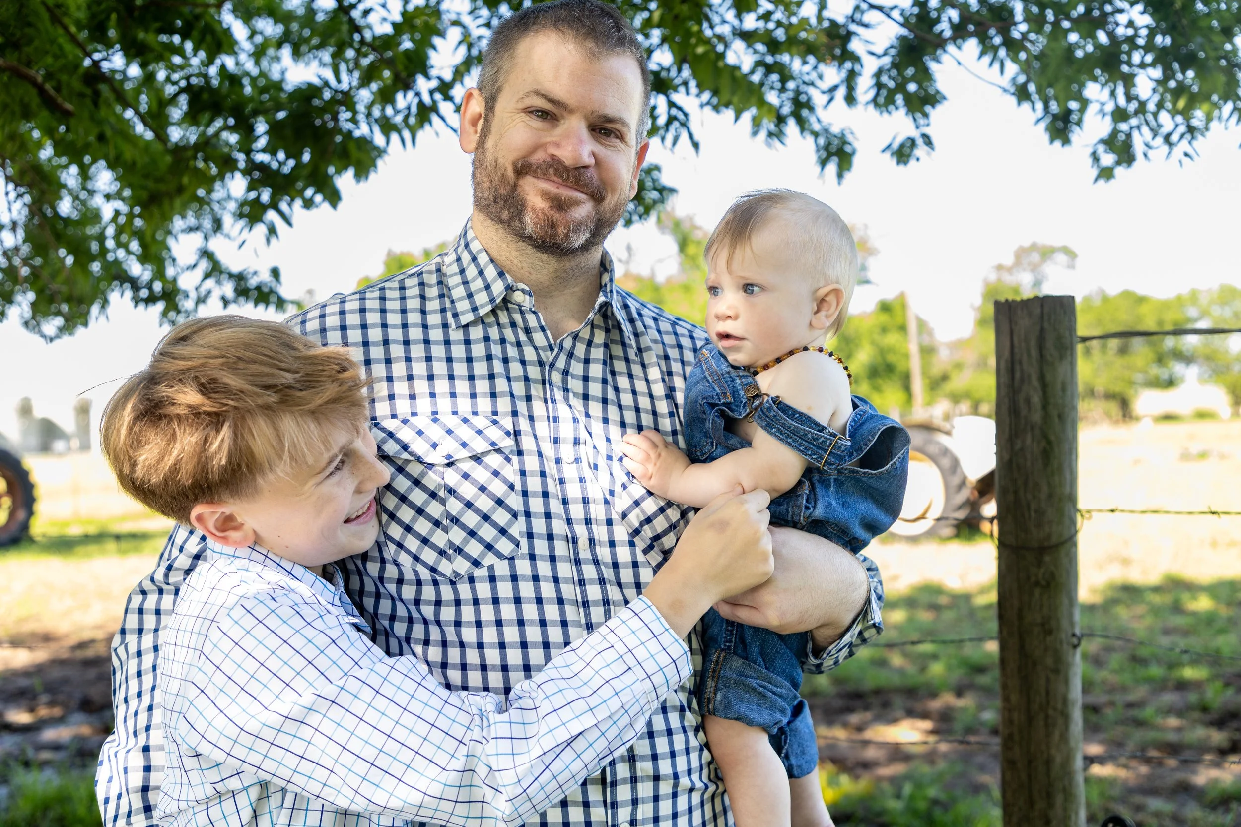 A man holding a young girl and a boy standing outside under a tree, with a farm fence in the background.