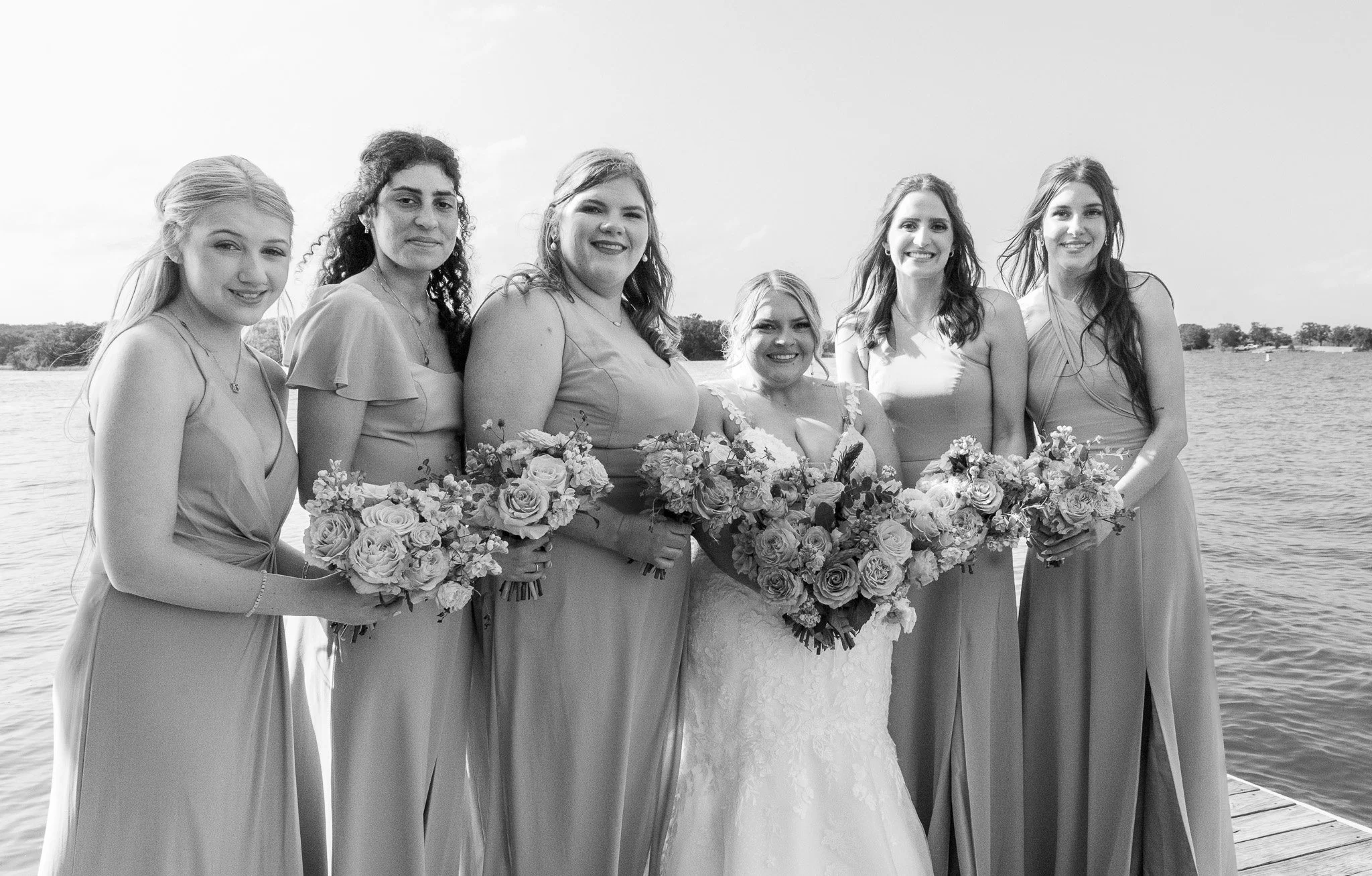Black and white photo of seven women standing on a dock by a lake, dressed in formal gowns, holding bouquets of flowers, smiling at the camera.