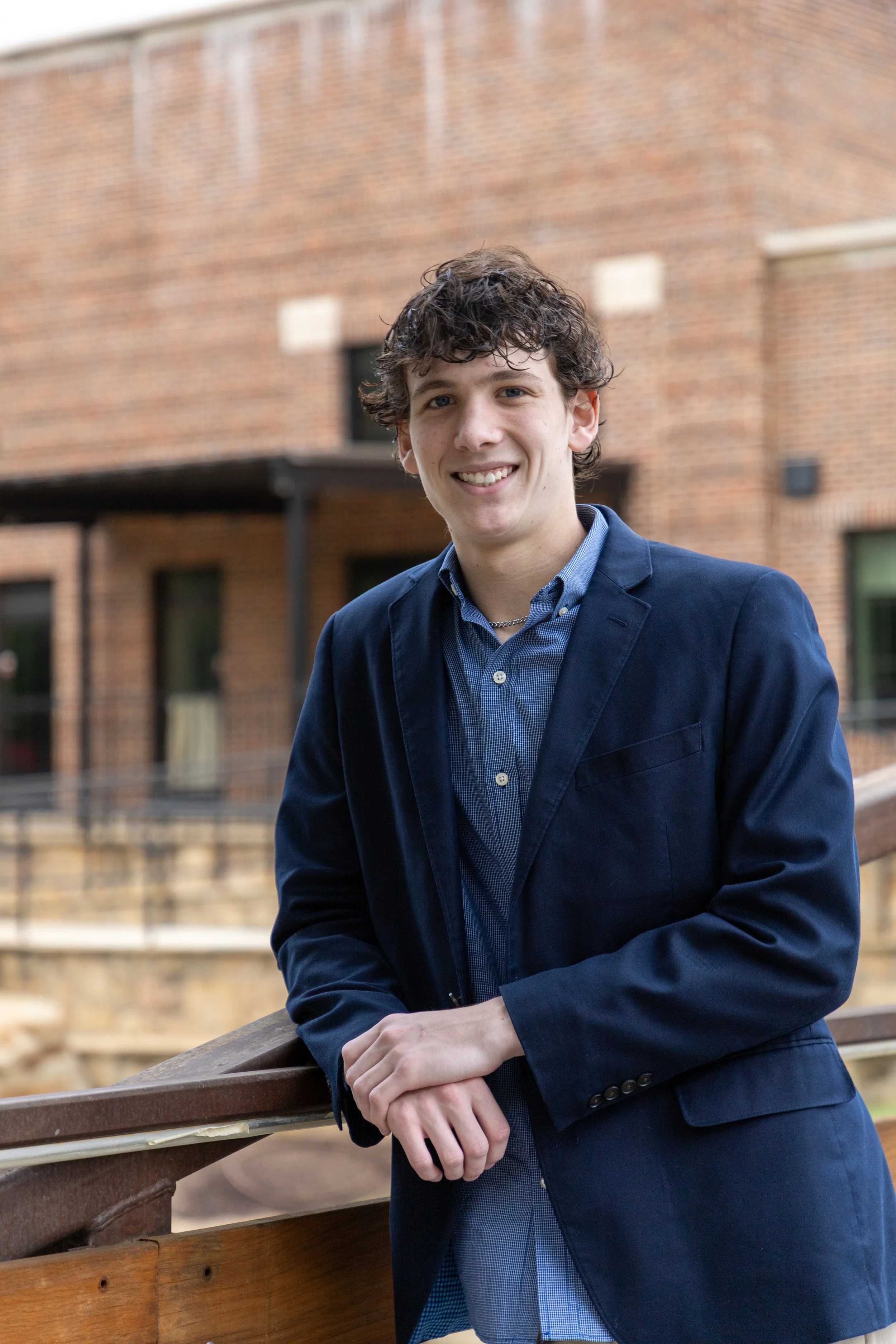 A young man with curly brown hair, dressed in a dark blue blazer and blue shirt, is leaning on a wooden railing outdoors in front of a brick building, smiling at the camera.