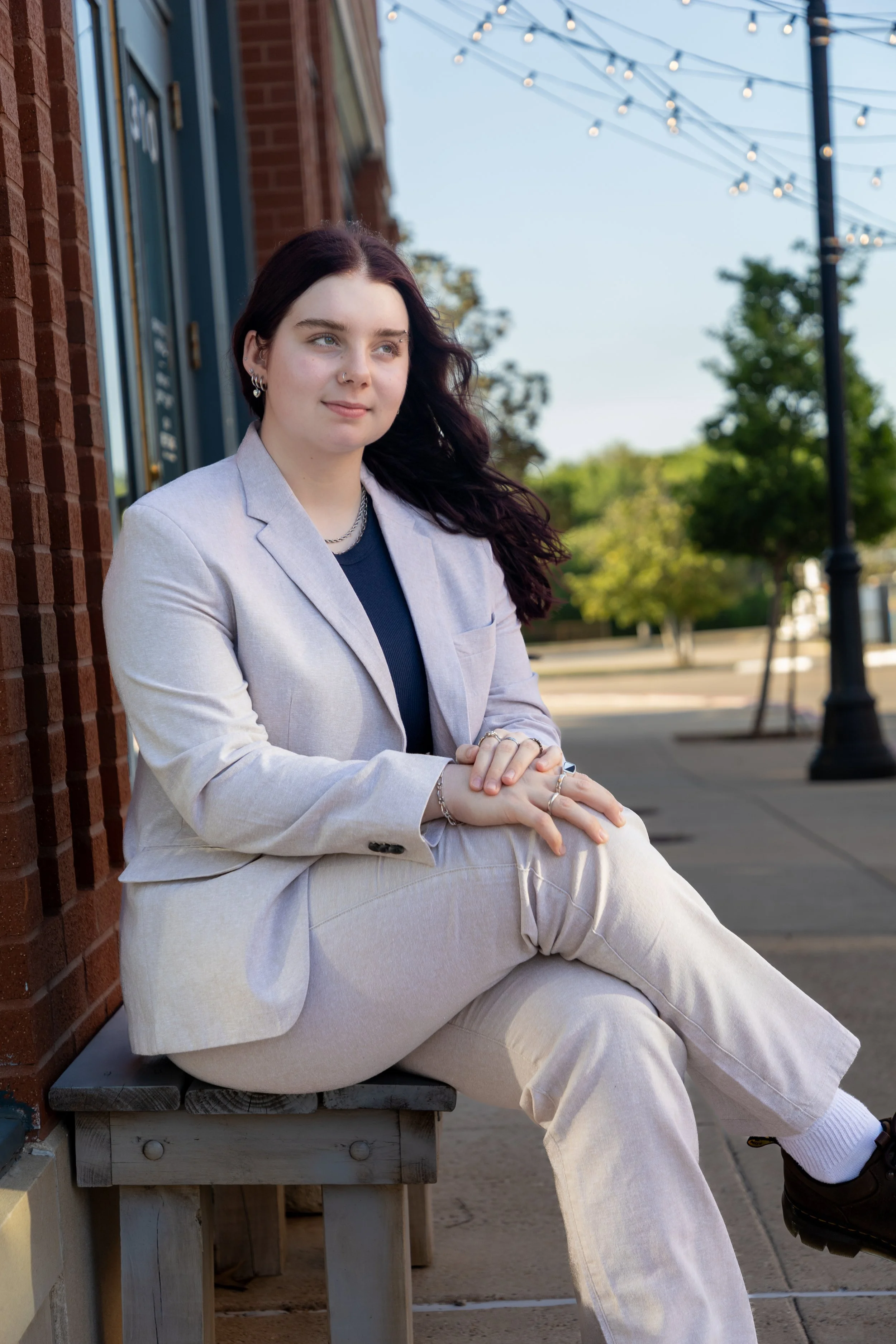 A young woman with dark hair sitting on a small wooden bench outside a brick building, wearing a light-colored suit and looking to the side, with string lights and trees in the background.