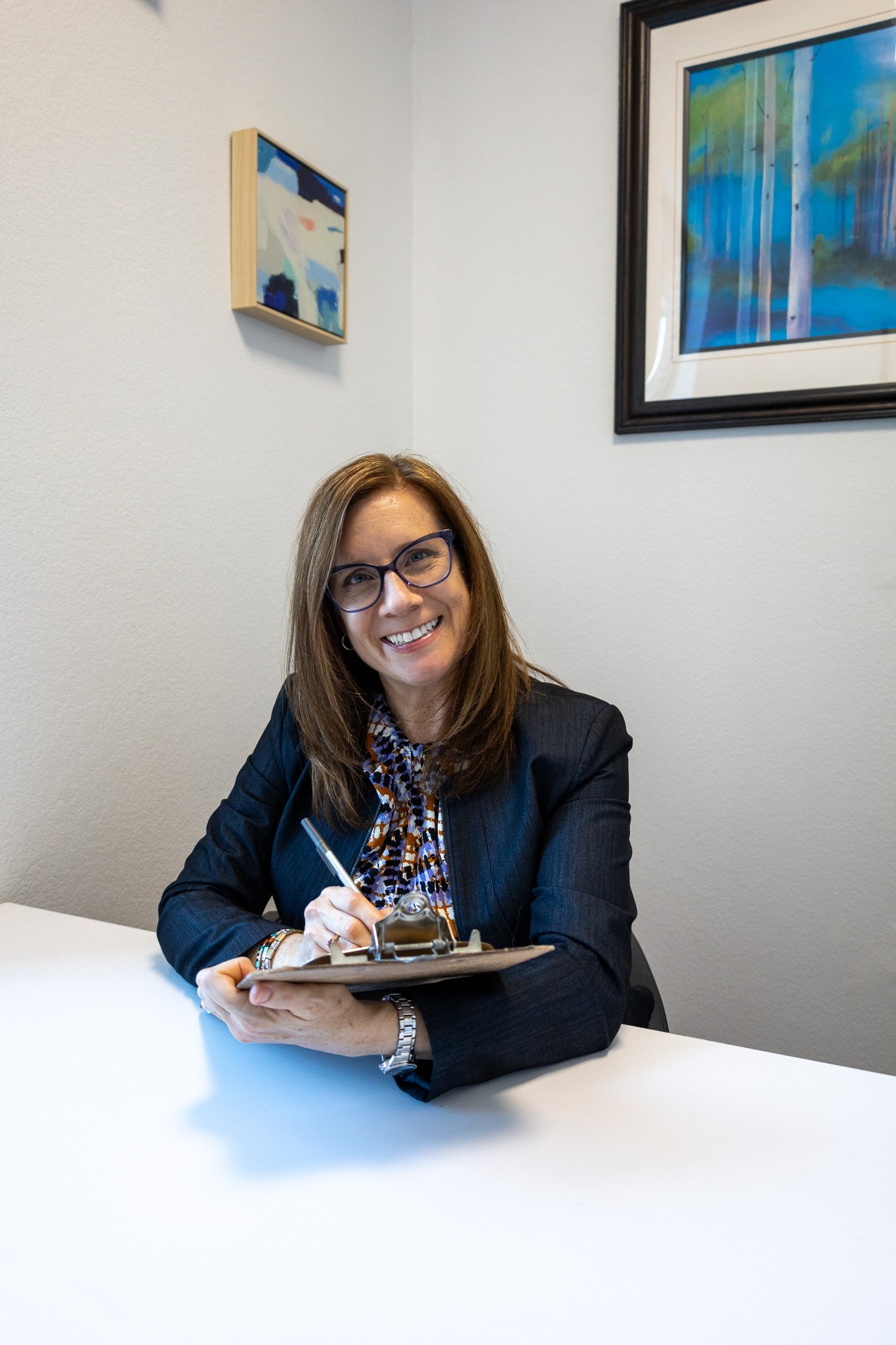 A woman with glasses smiling at camera, sitting at a desk with a clipboard, in a room with framed artwork on the wall.