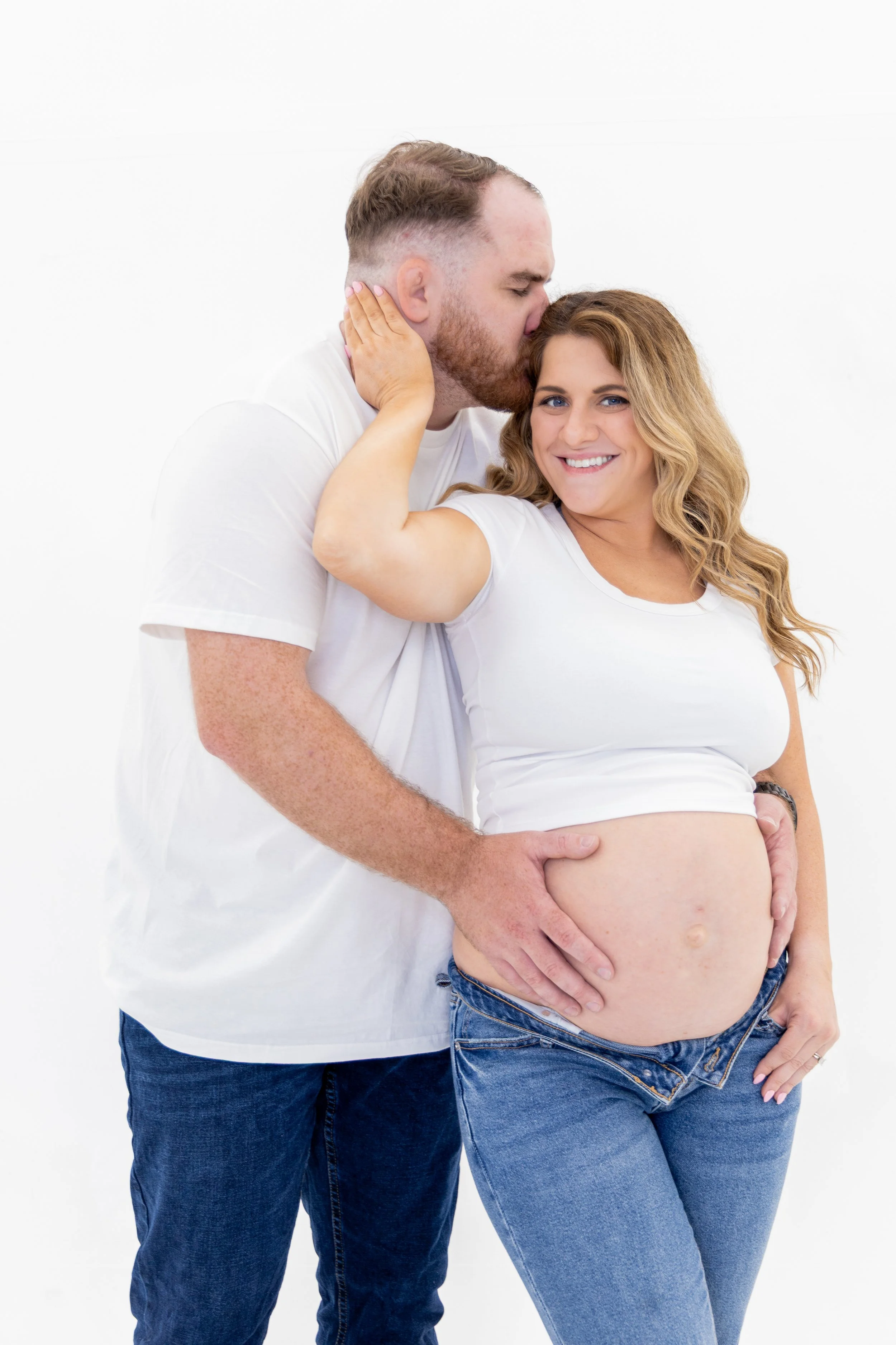 A pregnant woman with long wavy hair and blue eyes smiling, with her hand touching her hair, and a man with a beard and short hair kissing her forehead, both wearing white t-shirts and blue jeans against a white background.