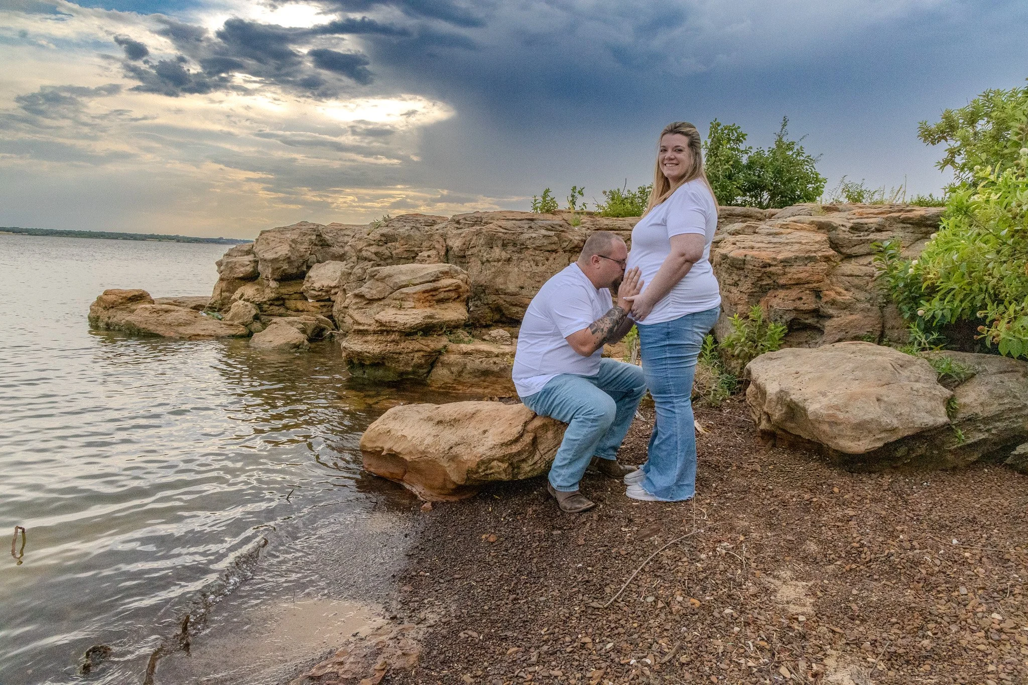 A couple stands on rocks near a lake, with one person kneeling and the other smiling at the camera. The sky is cloudy, and greenery is visible in the background.