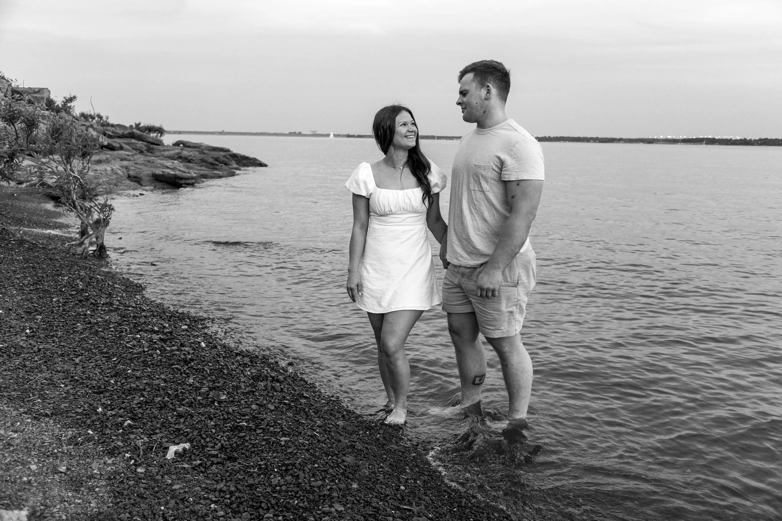 A smiling woman in a white dress and a man in a t-shirt and shorts holding hands in shallow water at the beach.