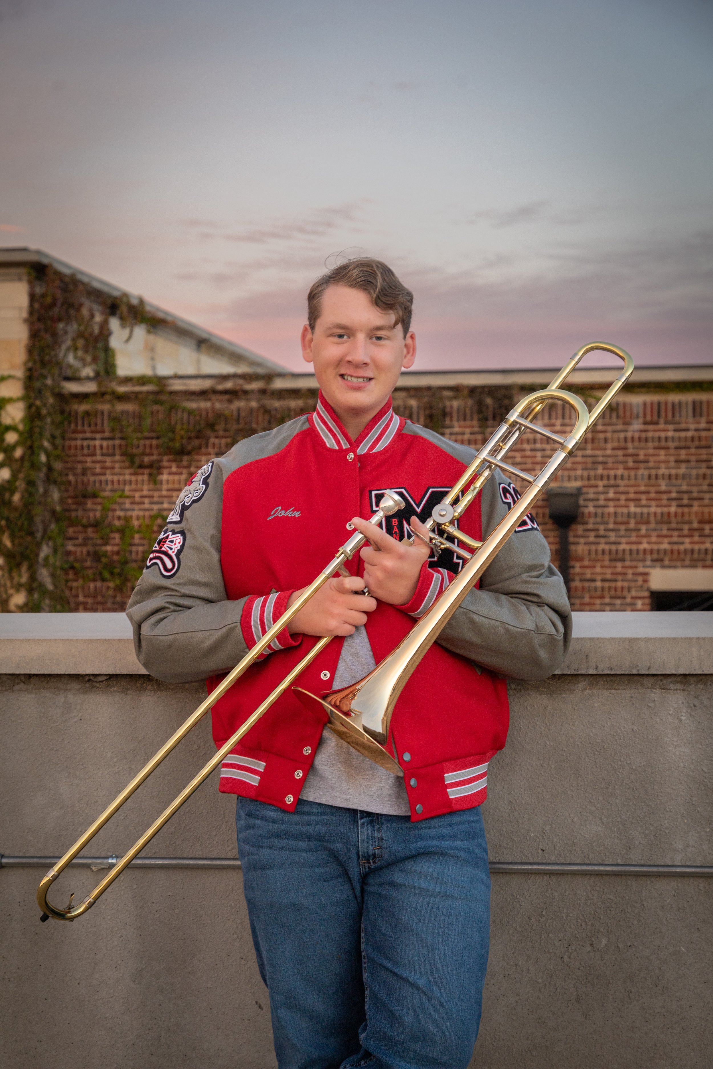 Young man in a red and gray letterman jacket holding a gold trombone outdoors during sunset.