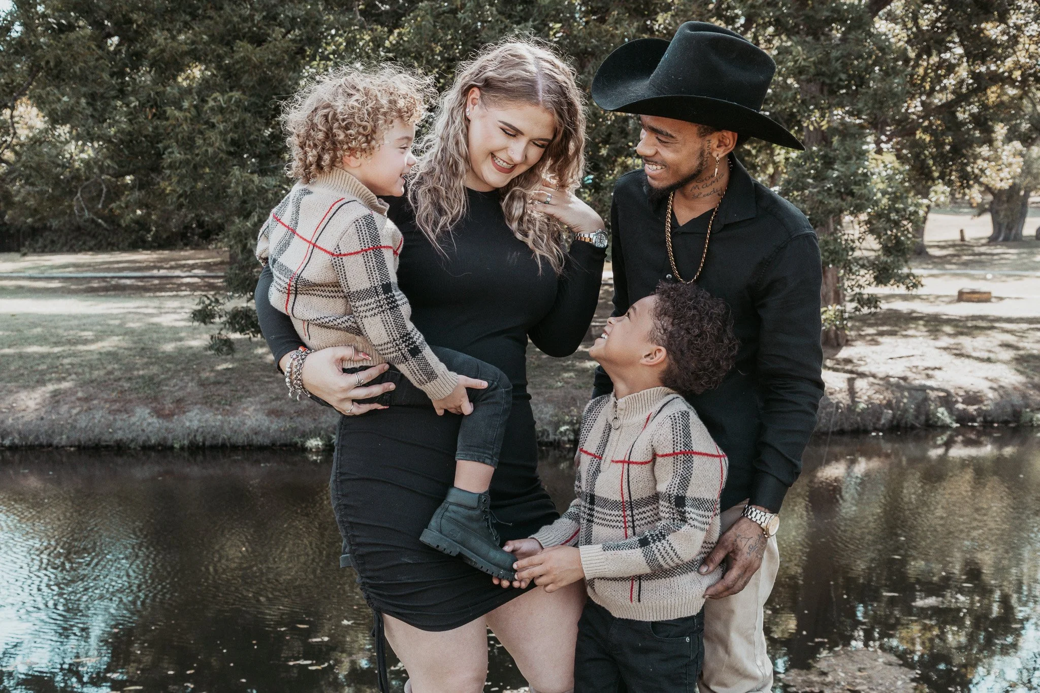 A diverse family enjoying a moment outdoors near a pond, with the mother holding one child in her arms, and the father and other child smiling at her, all dressed casually.