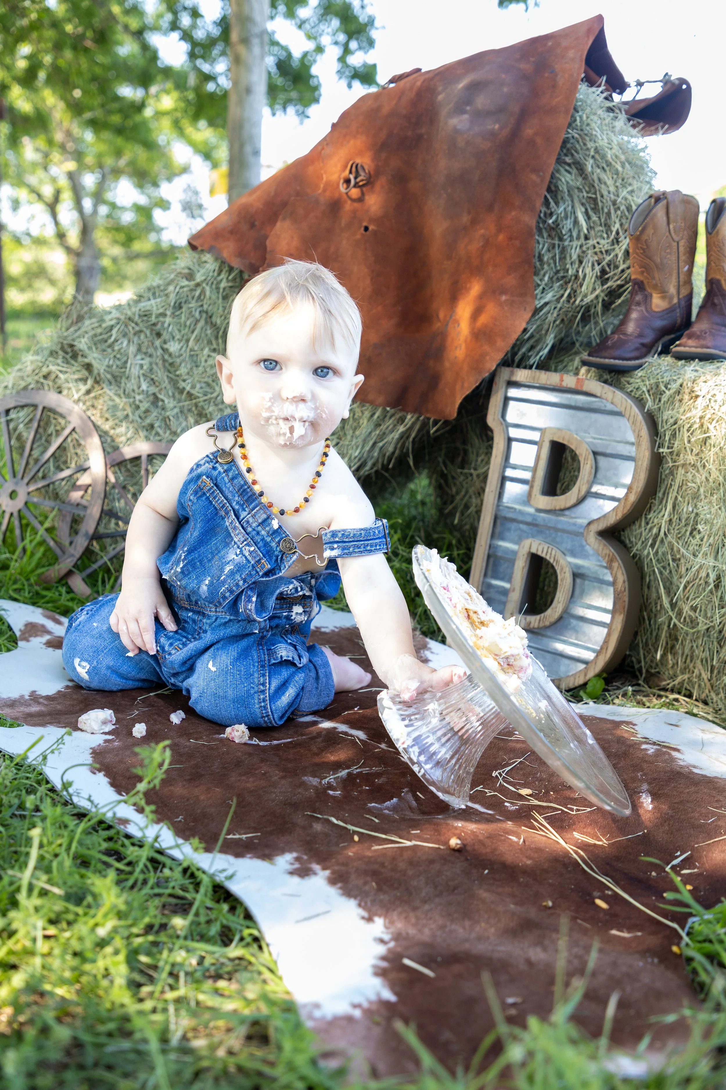 A young child with blonde hair and blue eyes, wearing denim overalls and a beaded necklace, sitting on a cowhide rug outdoors during the day, covered in cake and with cake on their face. There are hay bales, a metal 