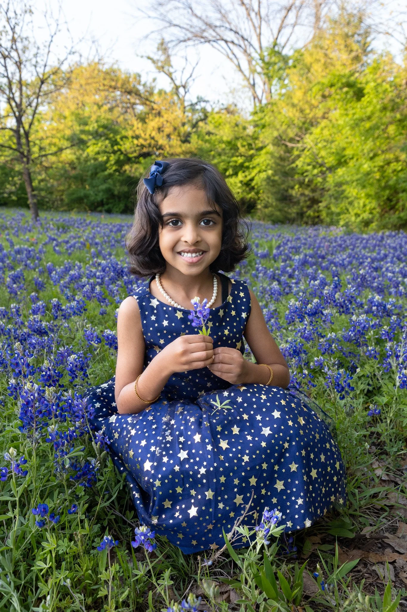 A young girl in a star-patterned navy dress sitting in a field of blue bonnets, holding a small bunch of blue flowers, with trees and a bright sky in the background.