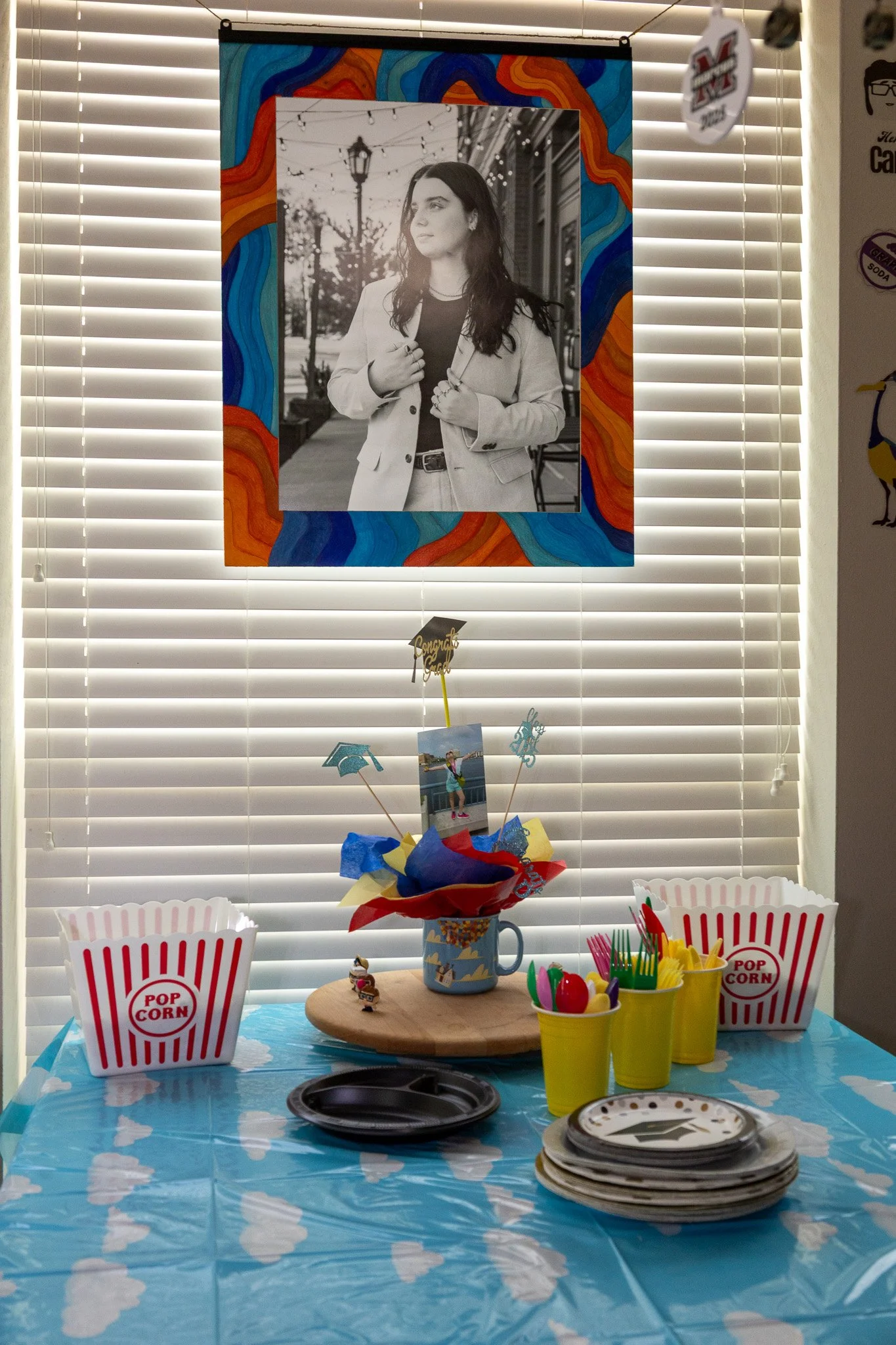 A decorated table set up for a celebration, with popcorn containers, colorful cups filled with plastic eggs, and a centerpiece with a graduation photo and decorations. A black and white portrait of a woman is hanging on the window blinds behind the t