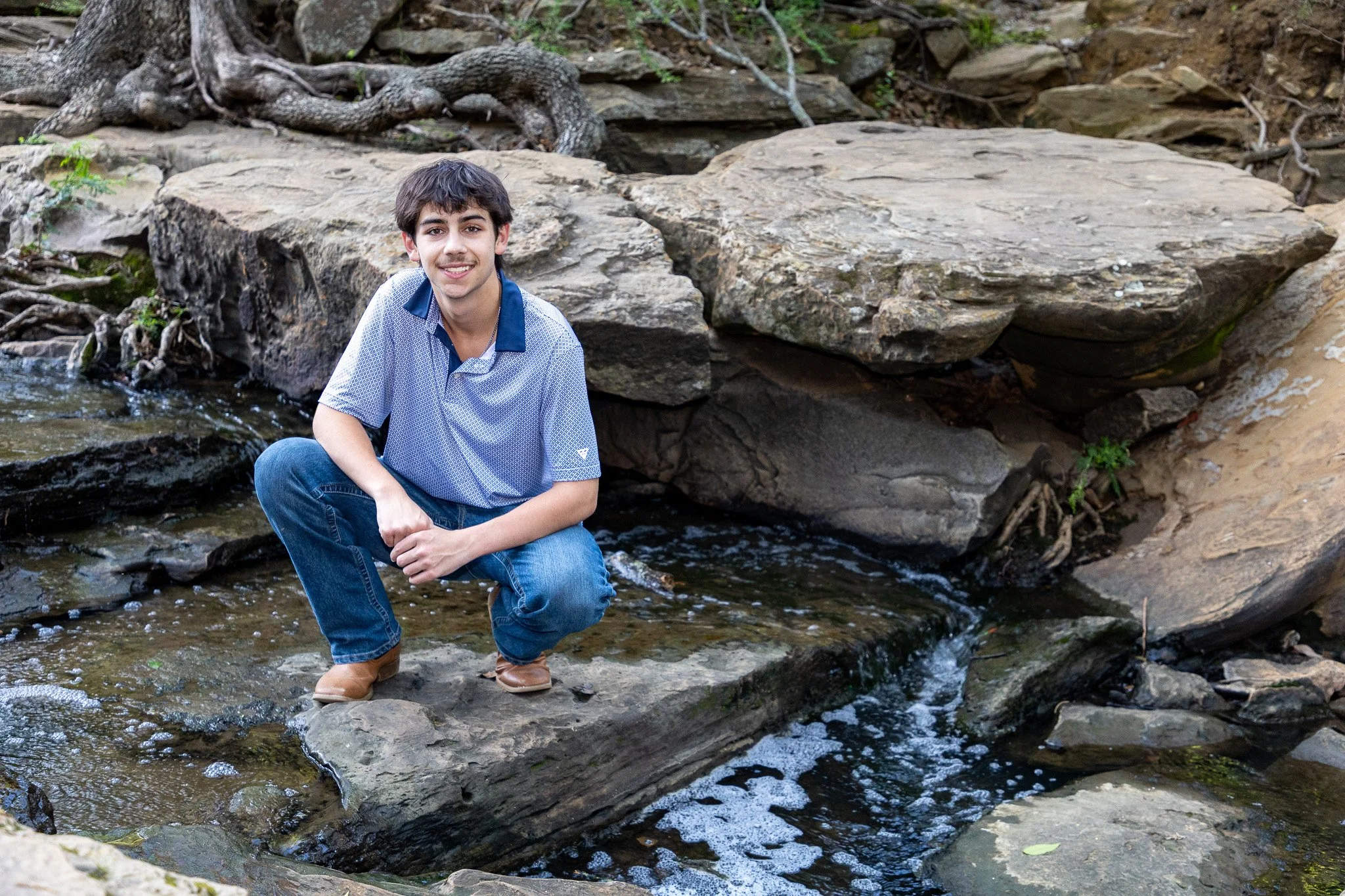 A young man squatting on a rock in a creek surrounded by large rocks and trees.