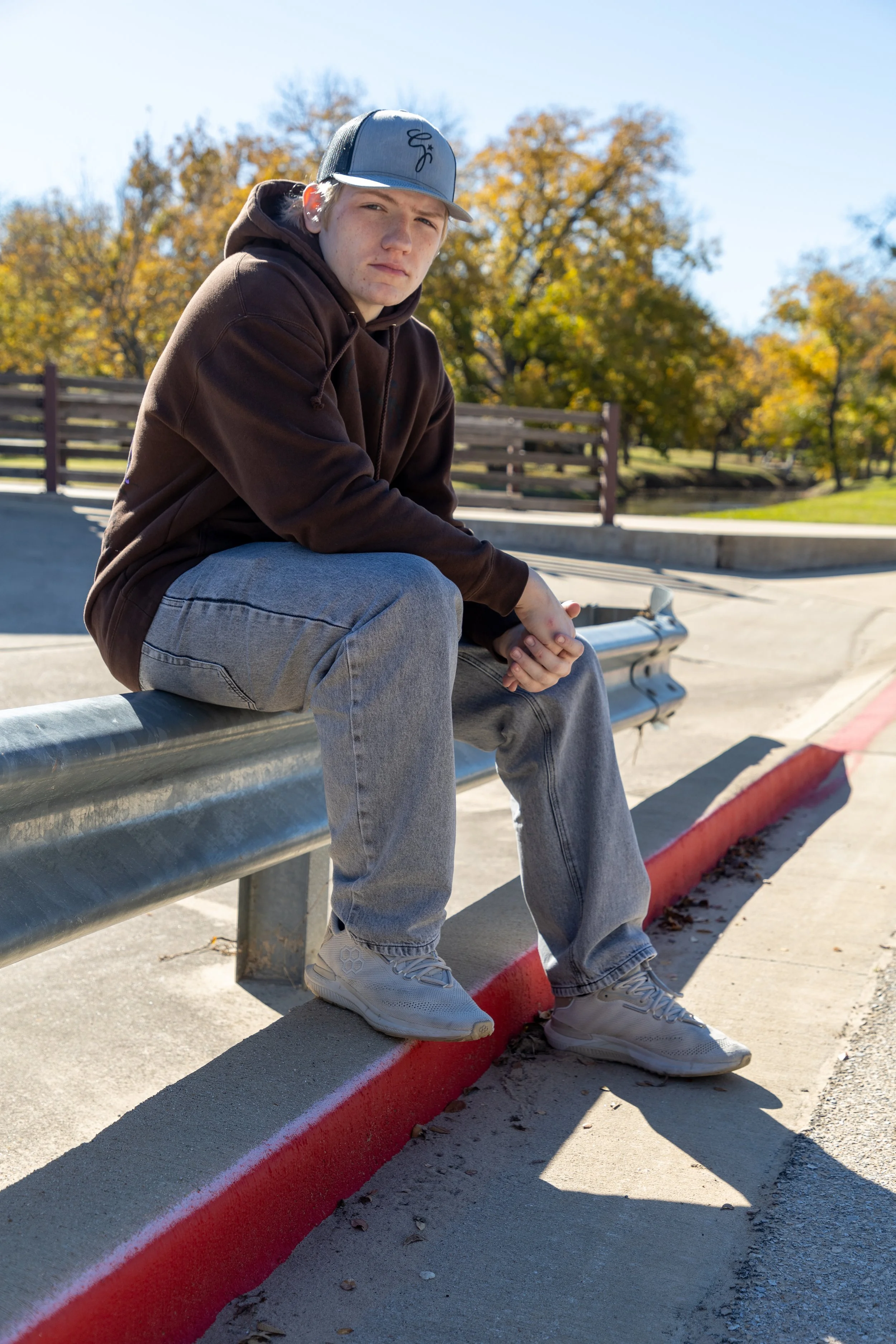A young man sitting on a concrete barrier at a skate park during autumn, wearing a brown hoodie, light gray jeans, white sneakers, and a baseball cap under a clear blue sky.