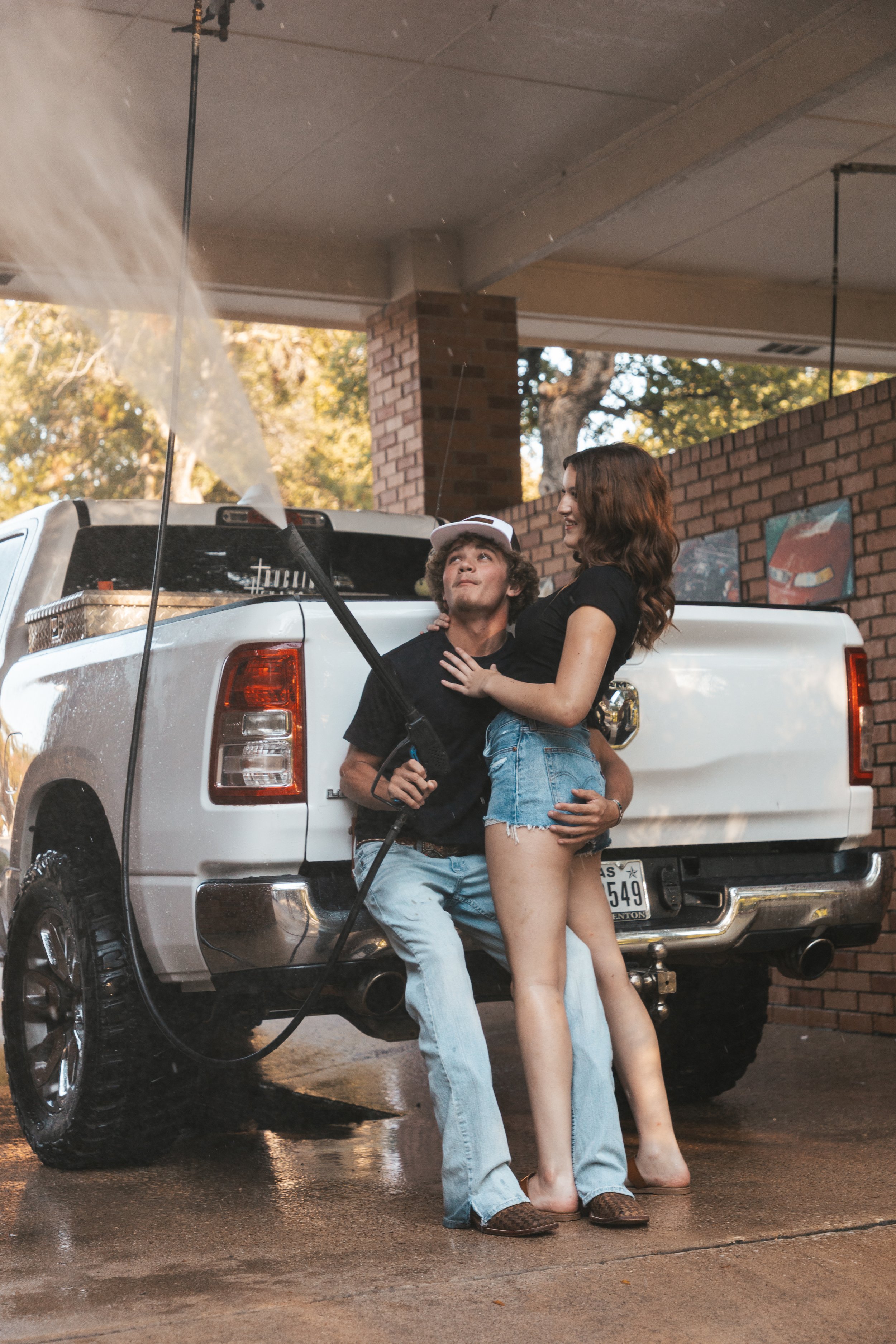 A young man and woman are at a car wash, with water and soap on a white pickup truck. The man is holding a hose, and they are smiling, sharing a joyful moment.