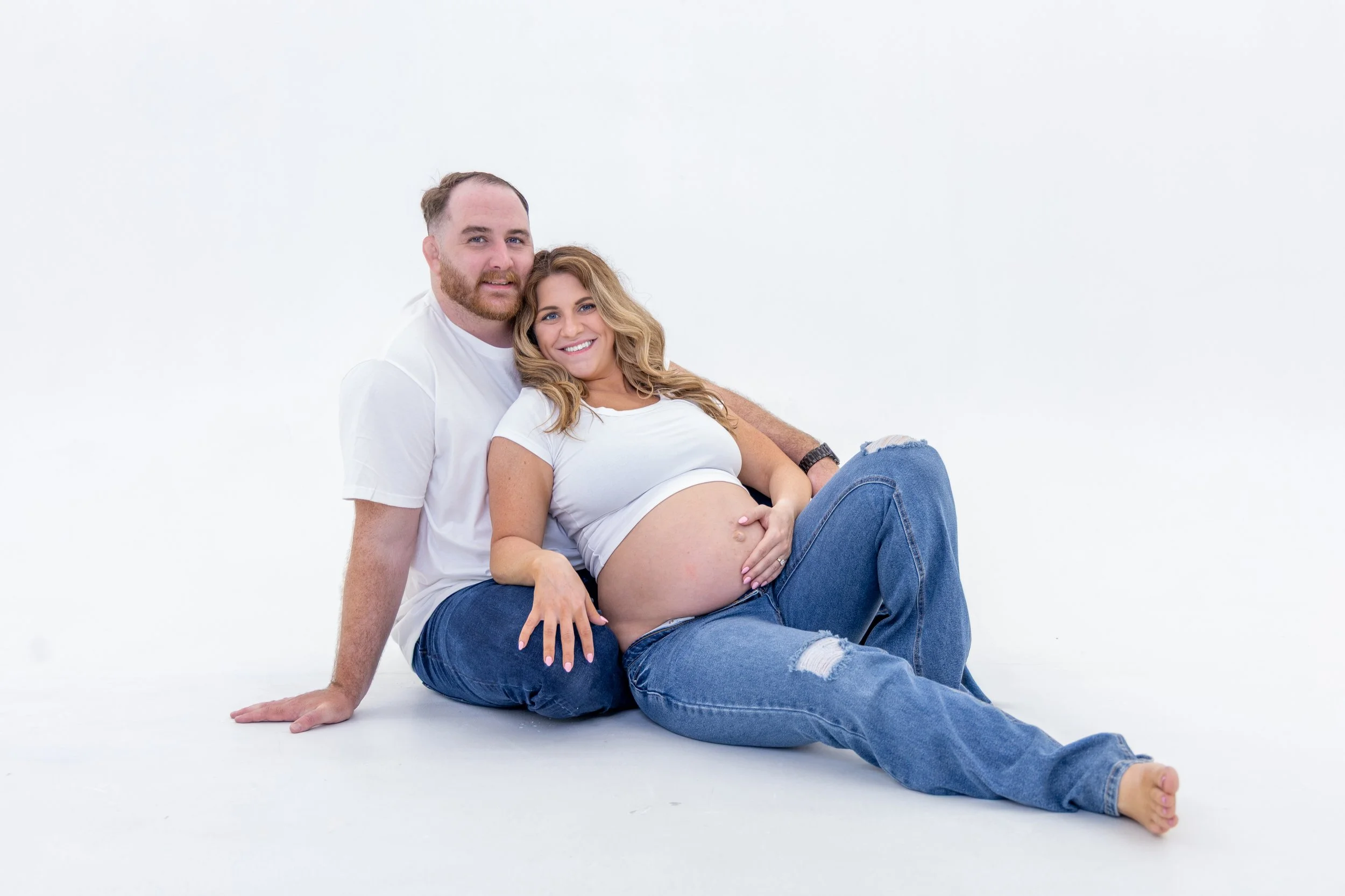 A pregnant woman sitting on the floor with her partner, both smiling, in a minimalistic white setting.