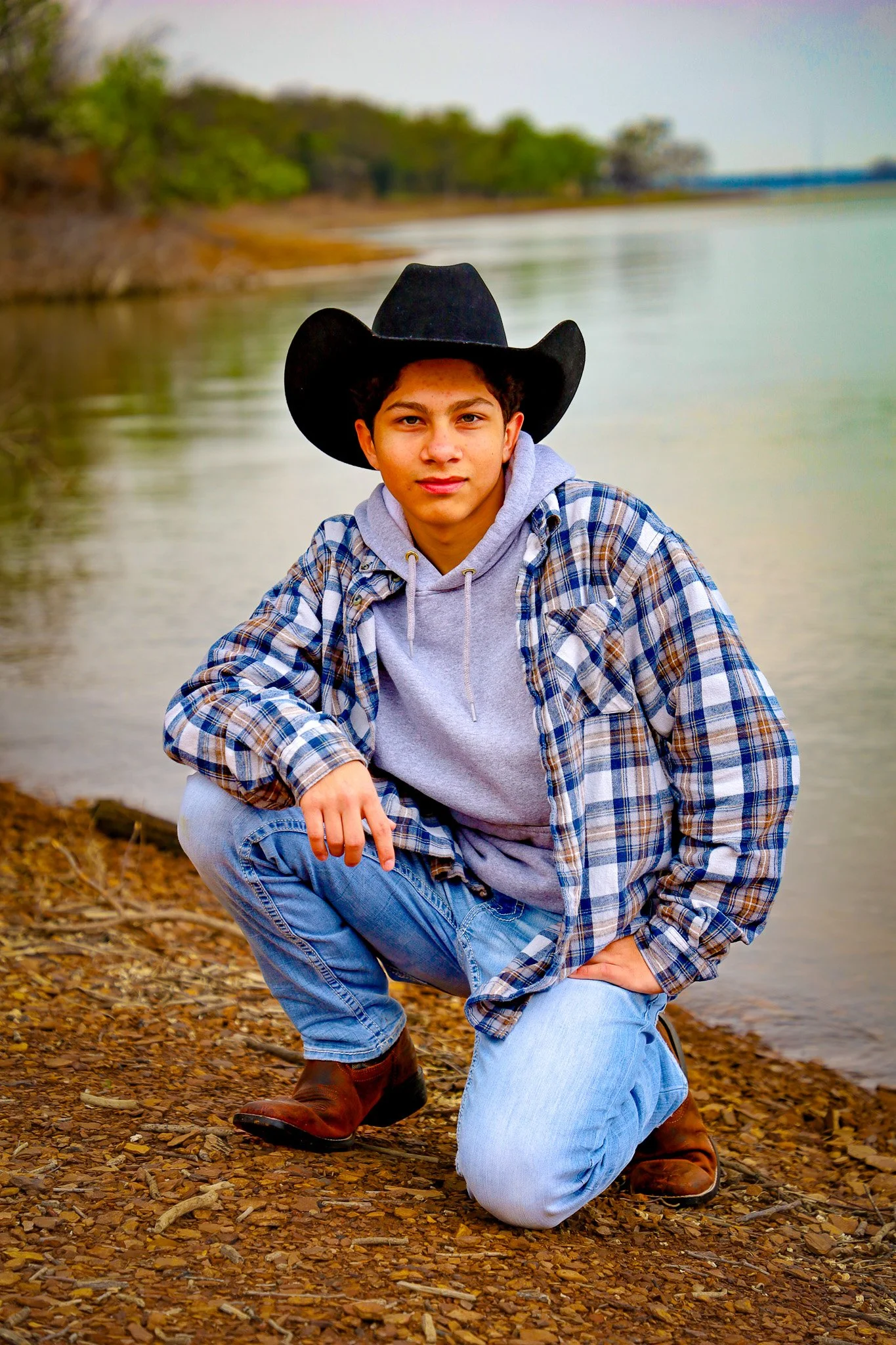 A young man wearing a black cowboy hat, gray hoodie, plaid jacket, and jeans squatting on a rocky shore by a body of water with a forested background.