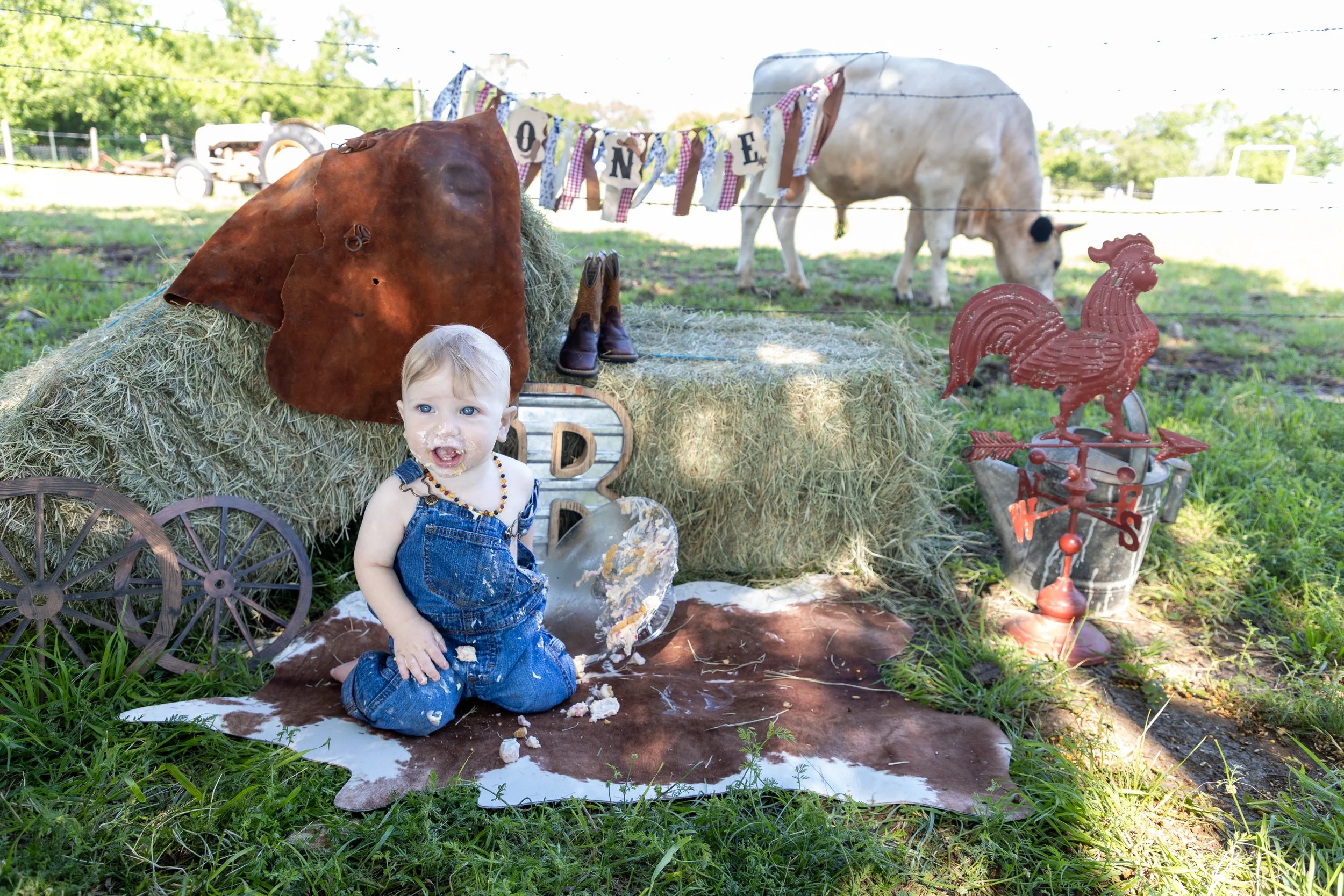 A young child with messy face and hands, wearing denim overalls and a beaded necklace, sitting on a cowhide rug outdoors surrounded by farm-themed decorations, including a hay bale, a rusted wagon wheel, a vintage milk can, a metal rooster weather va